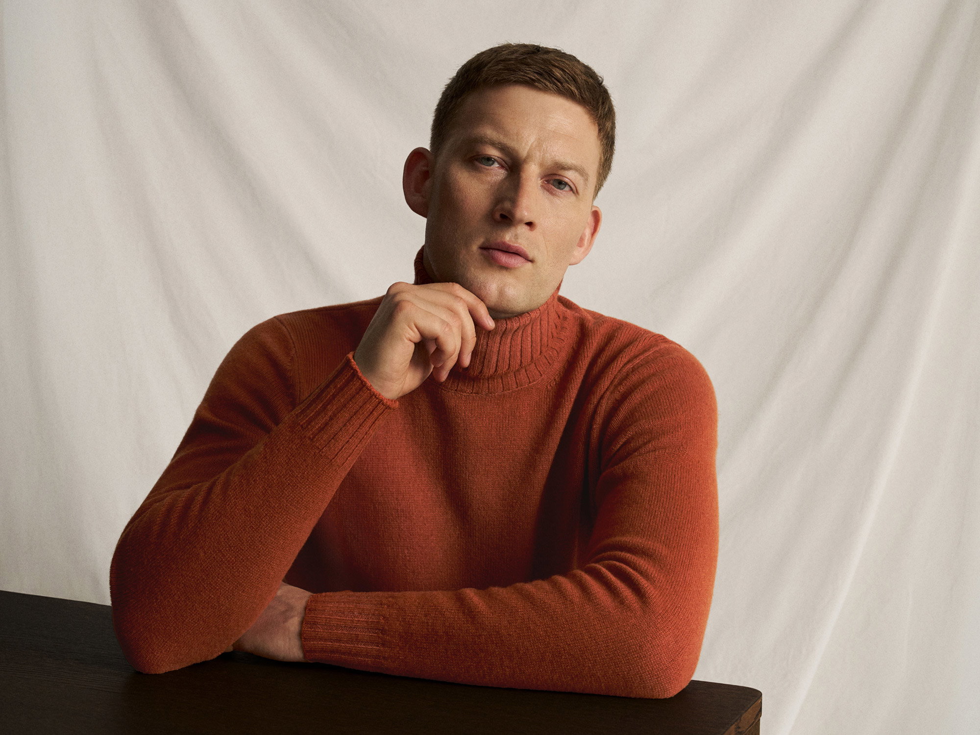 Portrait of a man with short brown hair wearing a rust-colored turtleneck sweater, resting his chin on his hand against a plain backdrop.