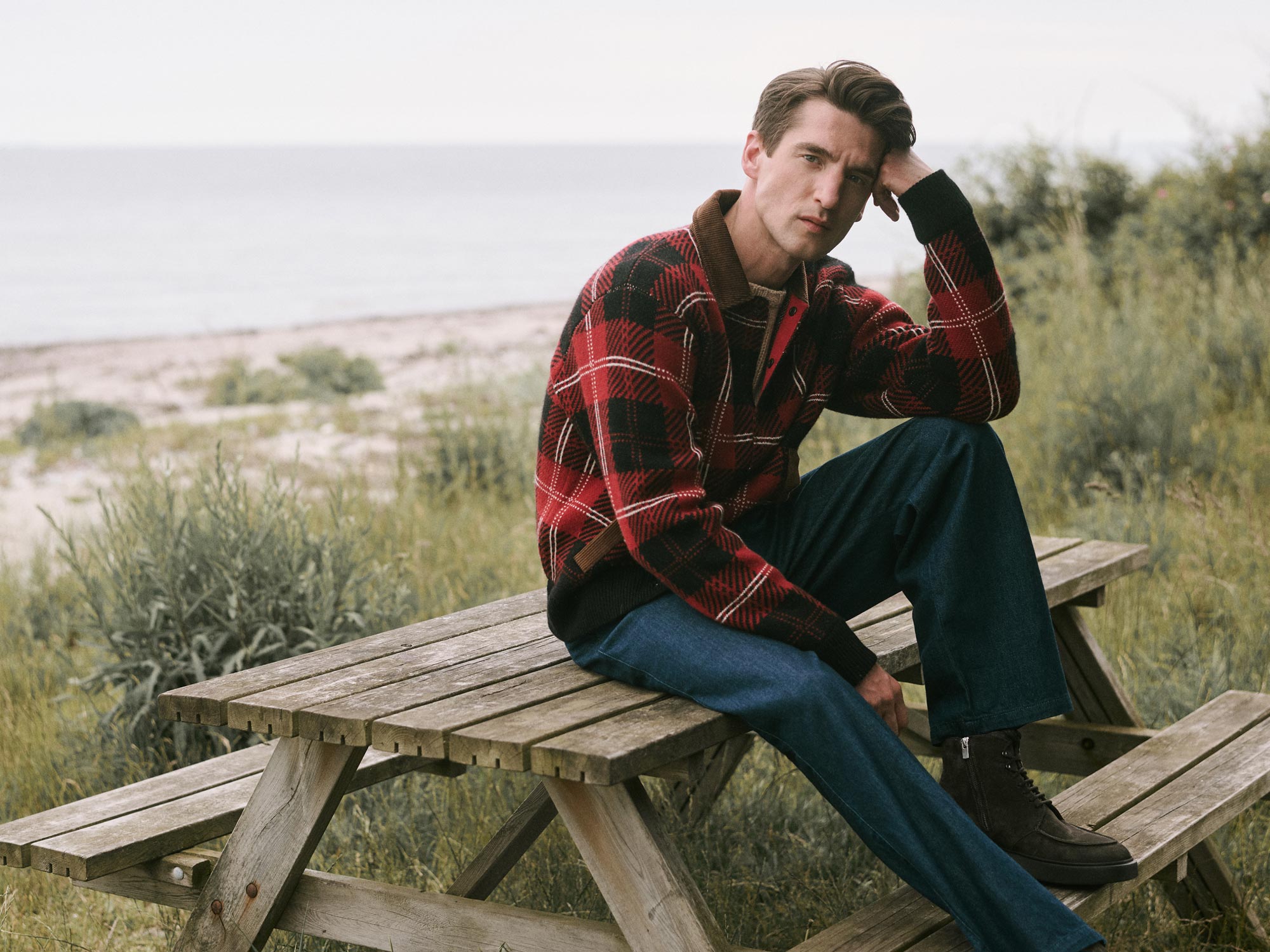 Mann in rotem Karo-Pullover und blauer Jeans sitzt nachdenklich auf einer Holzbank am Strand mit Meer und Grün im Hintergrund.