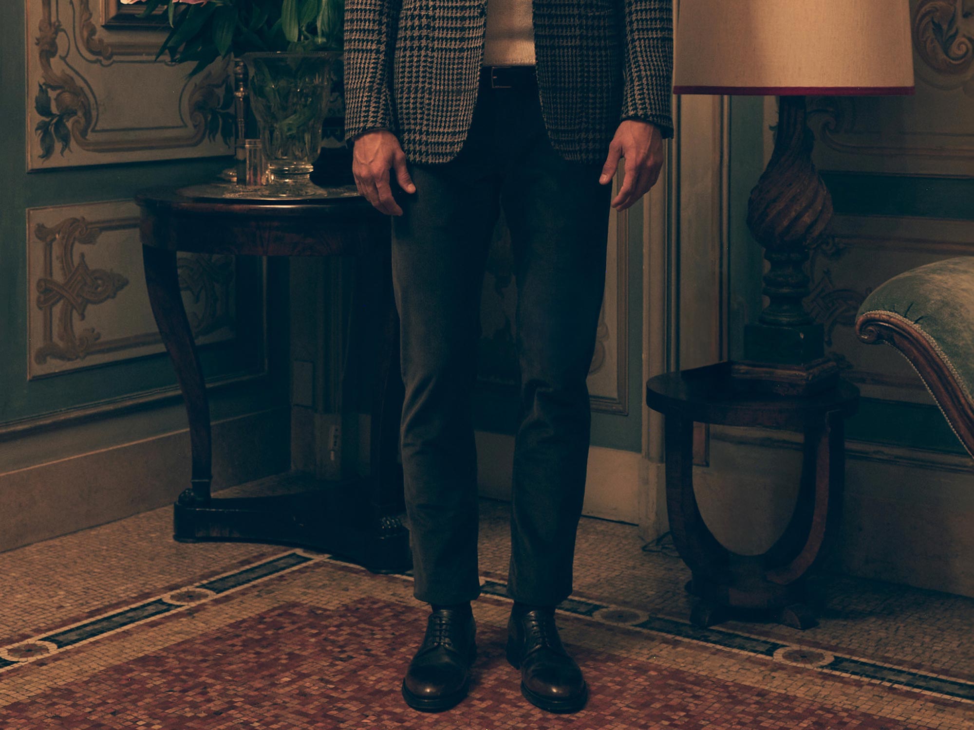 Mann trägt kariertes Sakko, weißen Pullover, graue Hose und schwarze Lederschuhe in elegant eingerichteten Raum.
