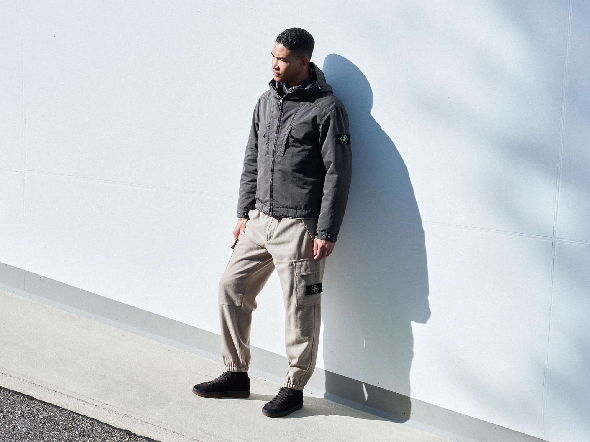 Junger Mann lehnt an weißer Wand, trägt graue Jacke mit Kapuze, beige Cargo-Hose und schwarze Sneaker, Schatten sichtbar.