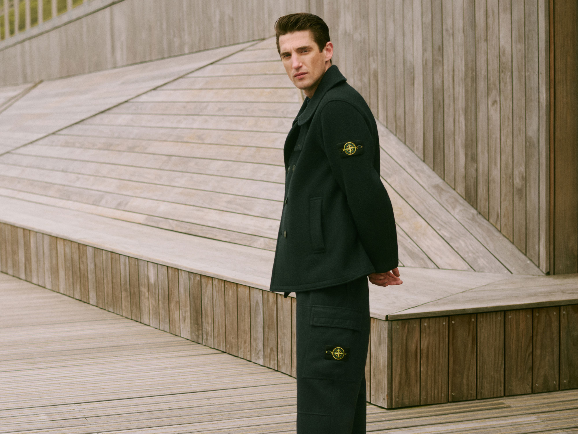Man in dark coat and matching pants with Stone Island logo patches standing against a wooden geometric wall background.