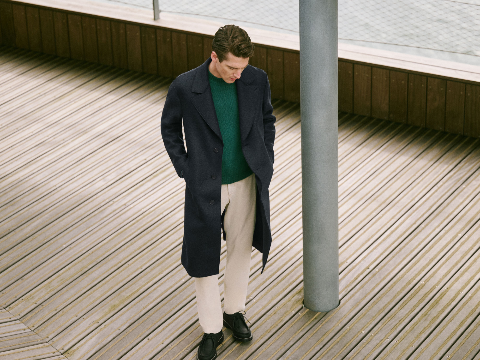 Man wearing a dark navy overcoat, green sweater, beige pants, and black shoes standing on a wooden deck next to a gray pole.