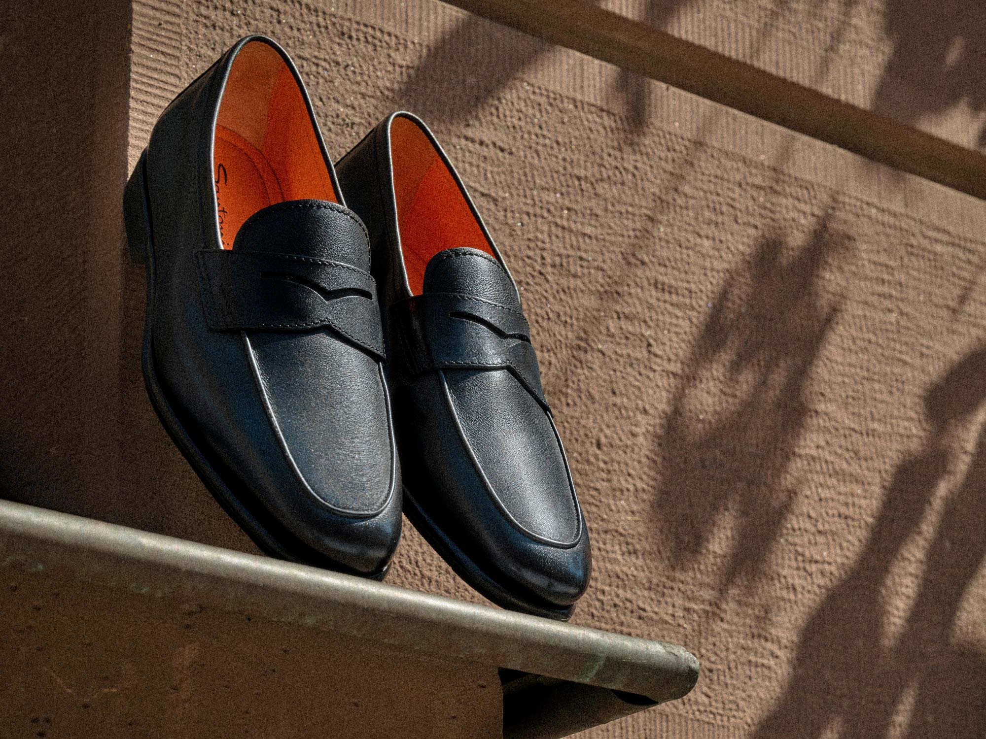 Black leather loafers with orange interior lining displayed on a concrete ledge against a textured brown wall.