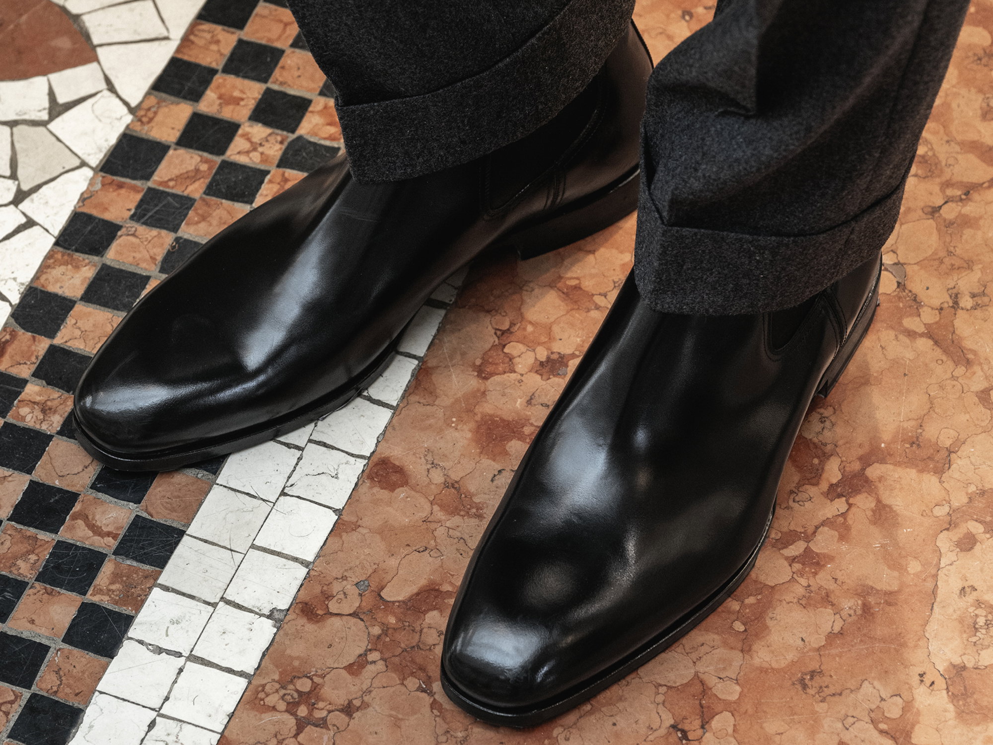 Schwarze, glänzende Chelsea-Boots aus Leder mit dunkelgrauer Hose auf gemustertem Marmorfußboden.