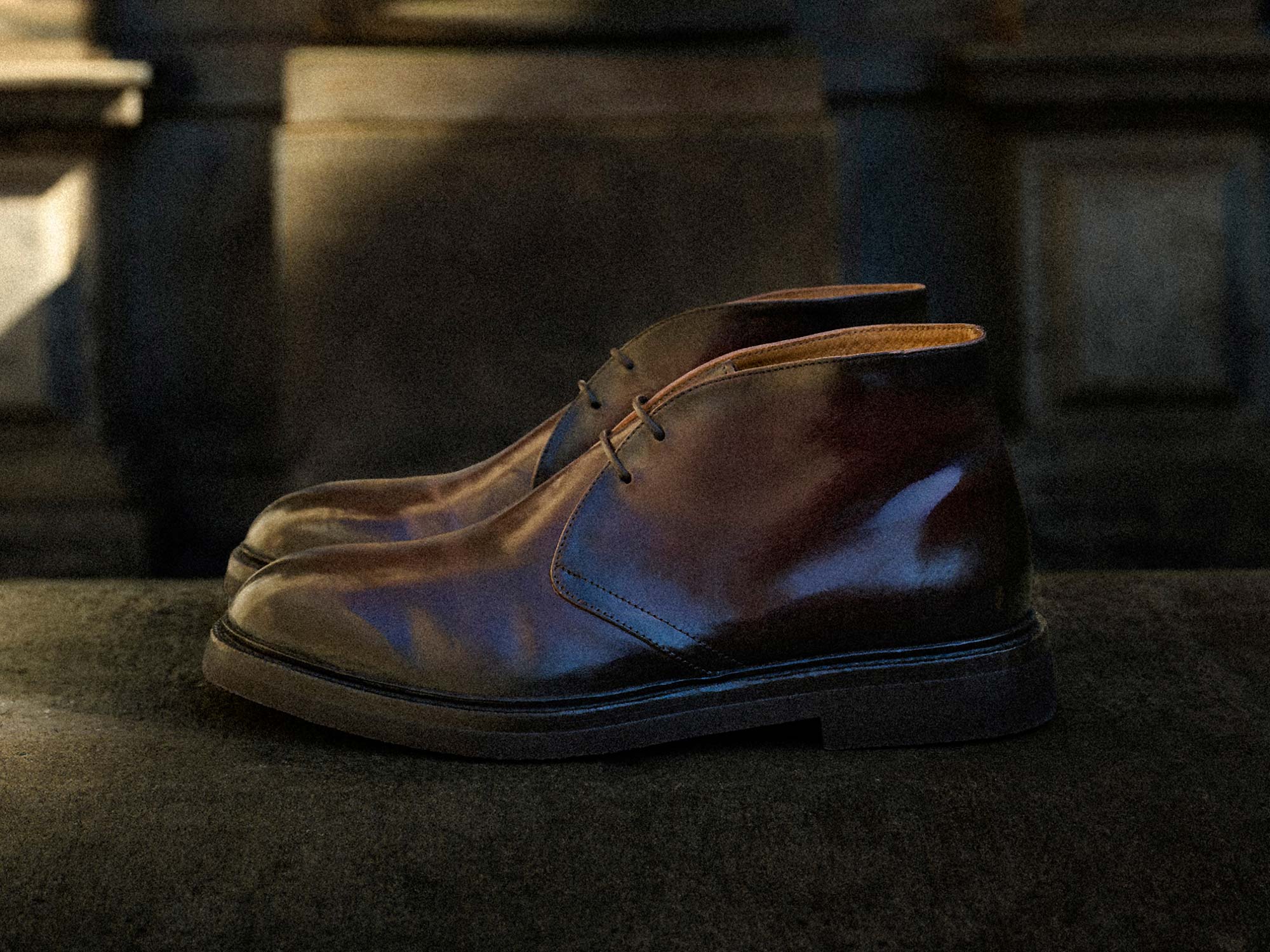 Brown leather ankle boots with laces, displayed on a stone surface against a dimly lit textured wall background.
