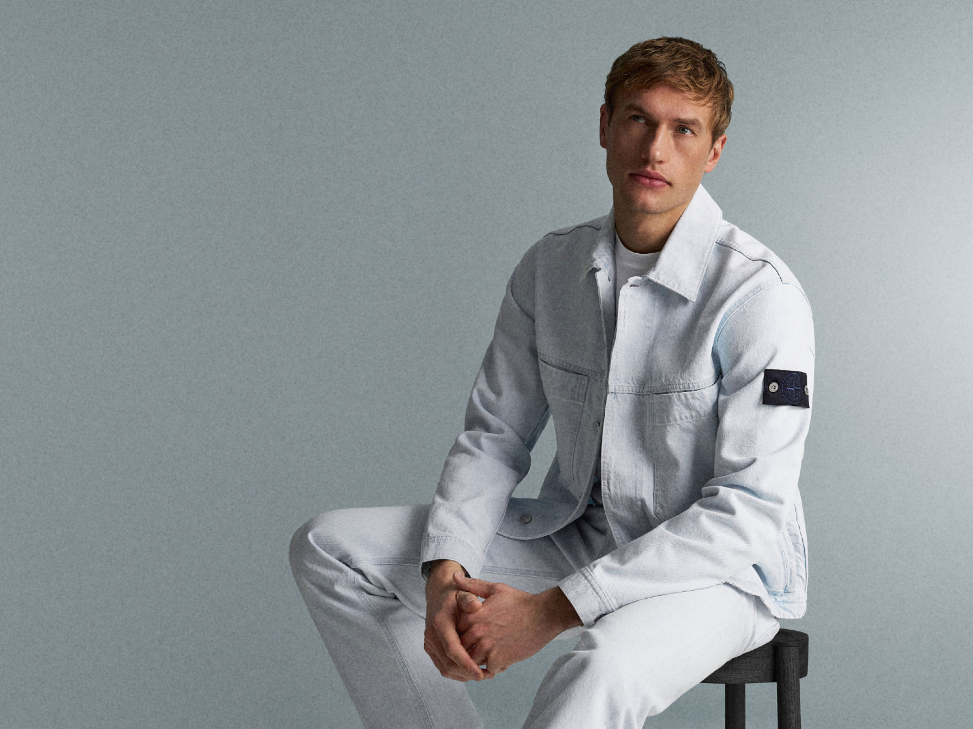 Young man with light brown hair wearing a light blue denim jacket and pants, sitting on a black stool against a gray background.