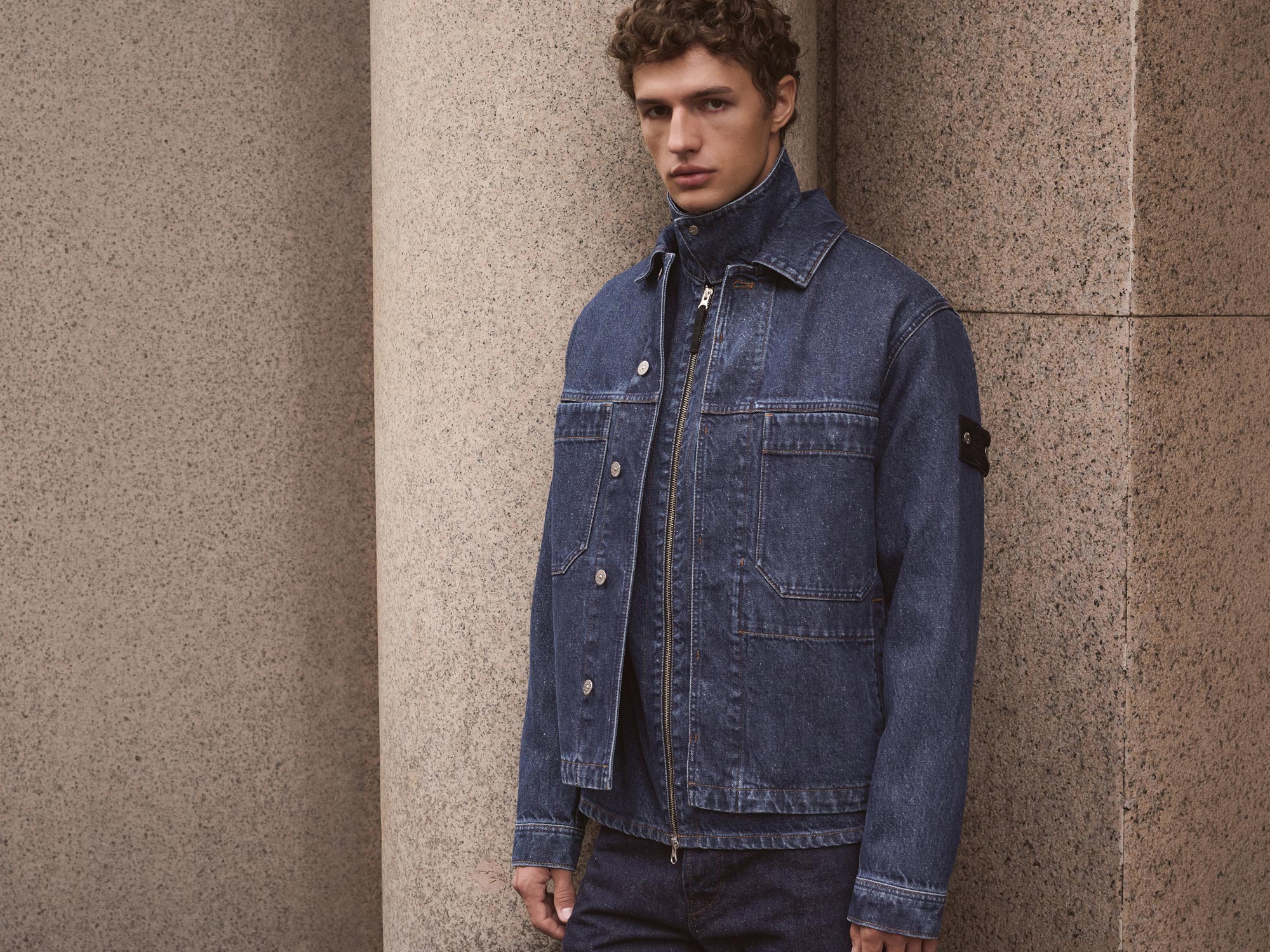 Junger Mann mit lockigem Haar trägt dunkelblaue Stone Island Denimjacke vor Granitsäulen.