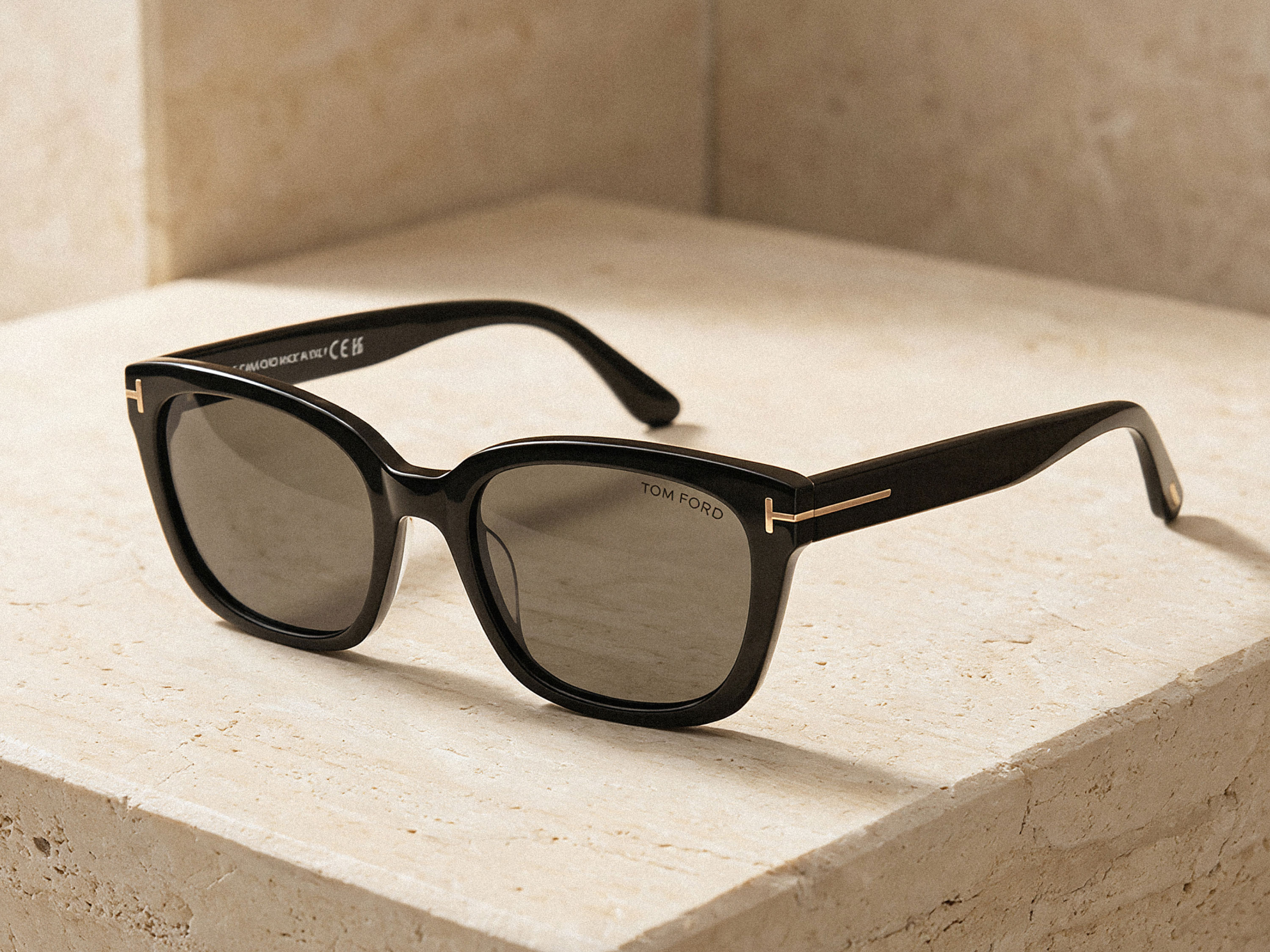 Schwarze Tom Ford Sonnenbrille mit breitem Rahmen und goldfarbenem T-Logo auf beigem Steinpodest vor beigem Hintergrund.
