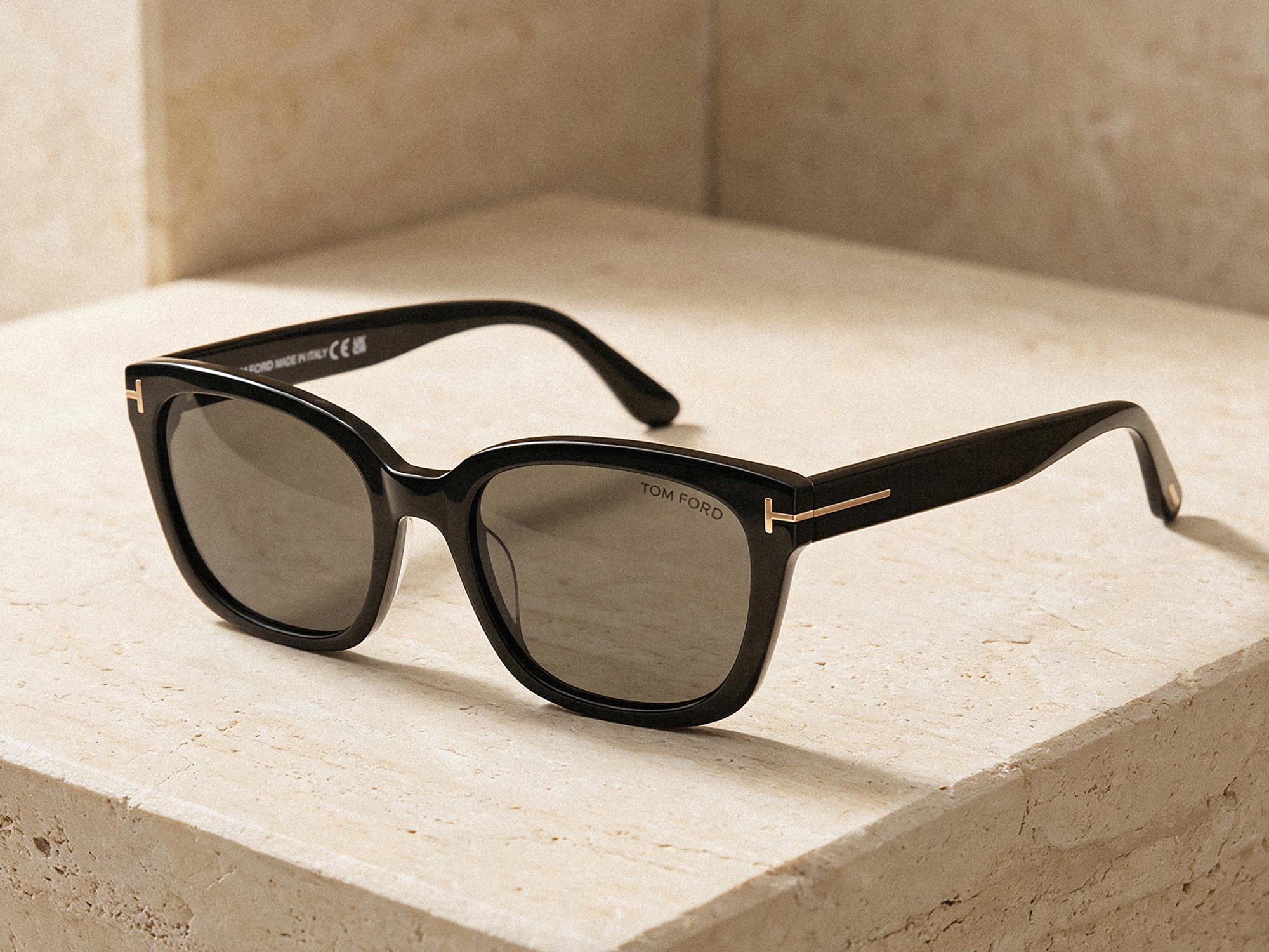 Black Tom Ford sunglasses with thick frames and dark lenses resting on a beige stone surface with gold accents on the temples.