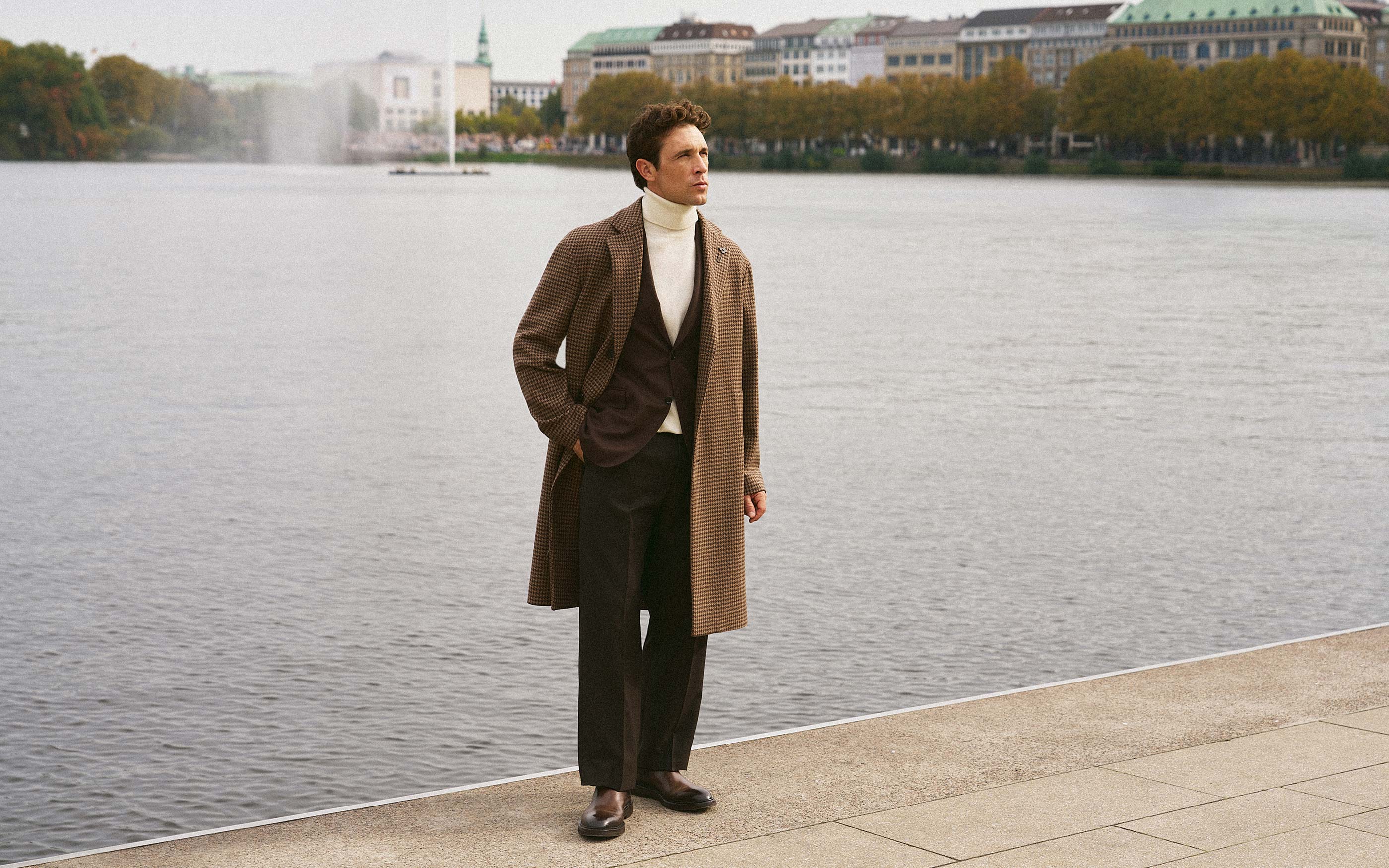 Man wearing a brown checkered overcoat, white turtleneck, black trousers, and brown shoes standing by a waterfront promenade.