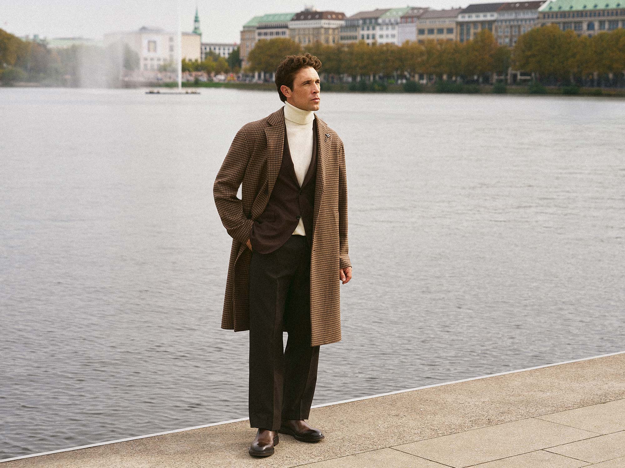 Man wearing a brown checkered long coat over a dark blazer and white turtleneck, standing by a lakeside promenade.