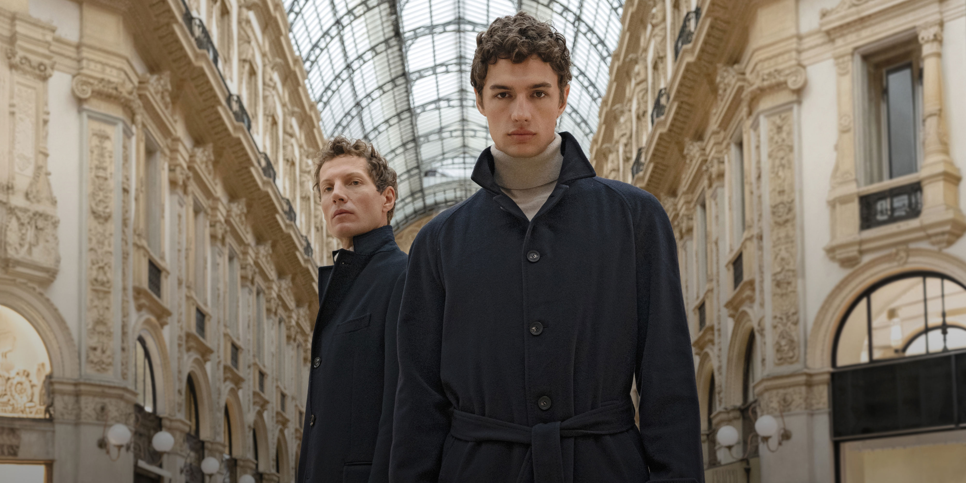 Two men wearing dark blue overcoats stand in an ornate shopping arcade with a glass ceiling and decorative walls.
