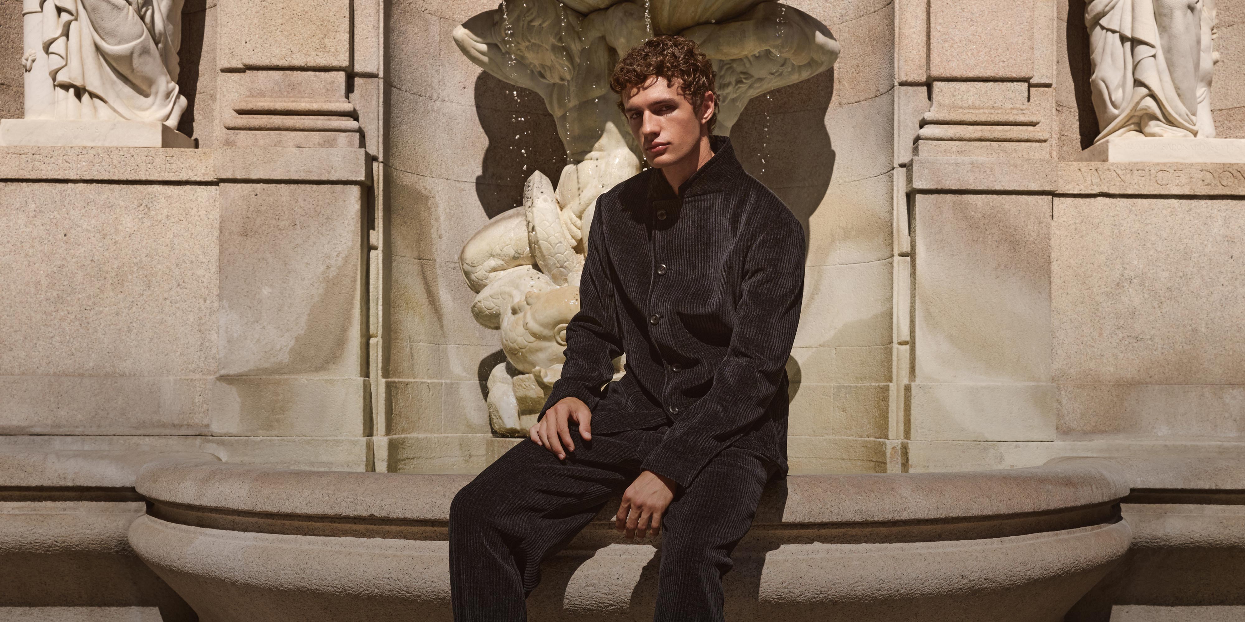 Young man with curly hair wearing a dark corduroy jacket and pants sitting on a stone fountain with sculpted figures.