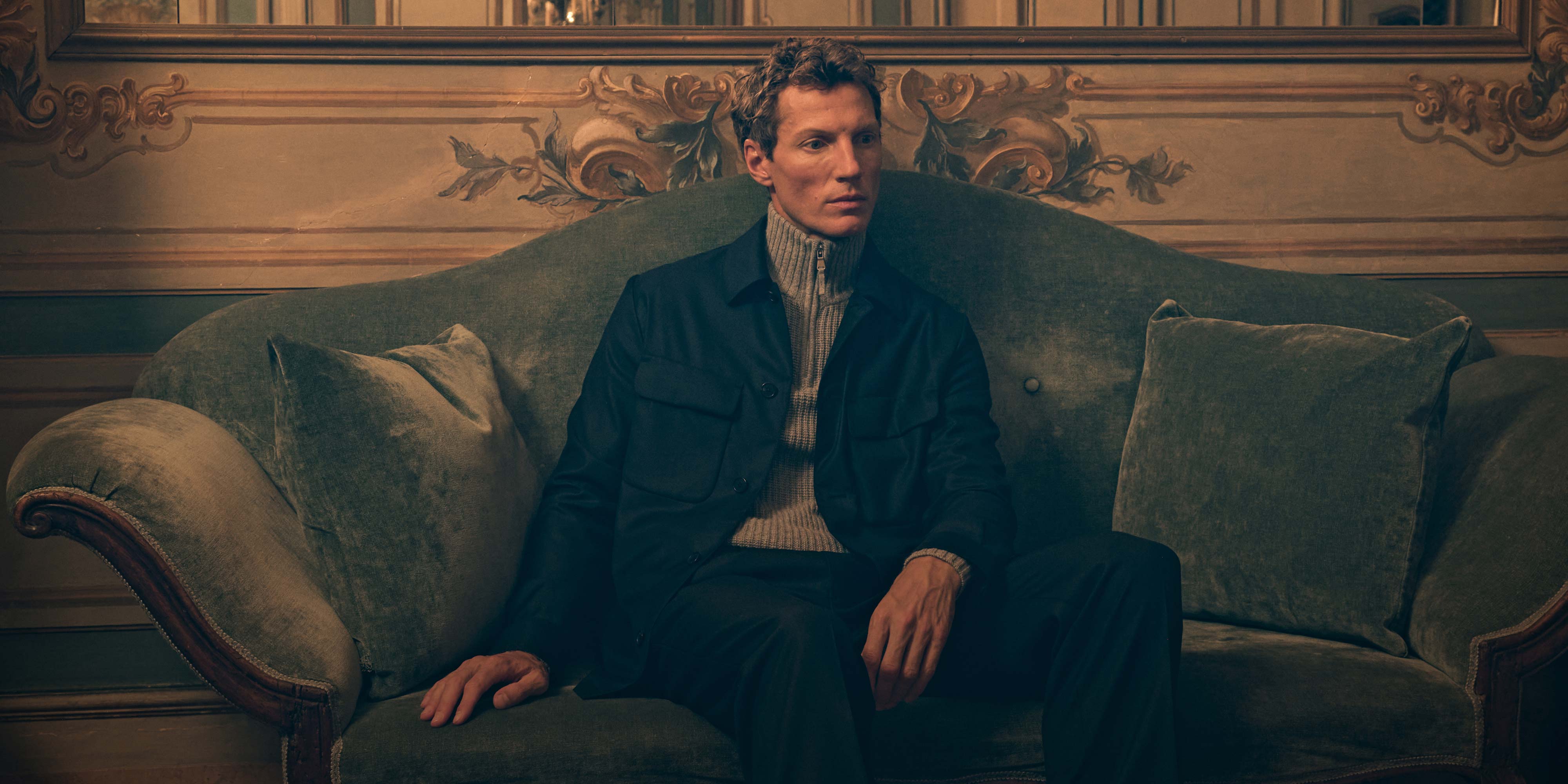 Man wearing a dark jacket and beige turtleneck sweater sitting on an olive green vintage sofa with ornate wall decor behind.