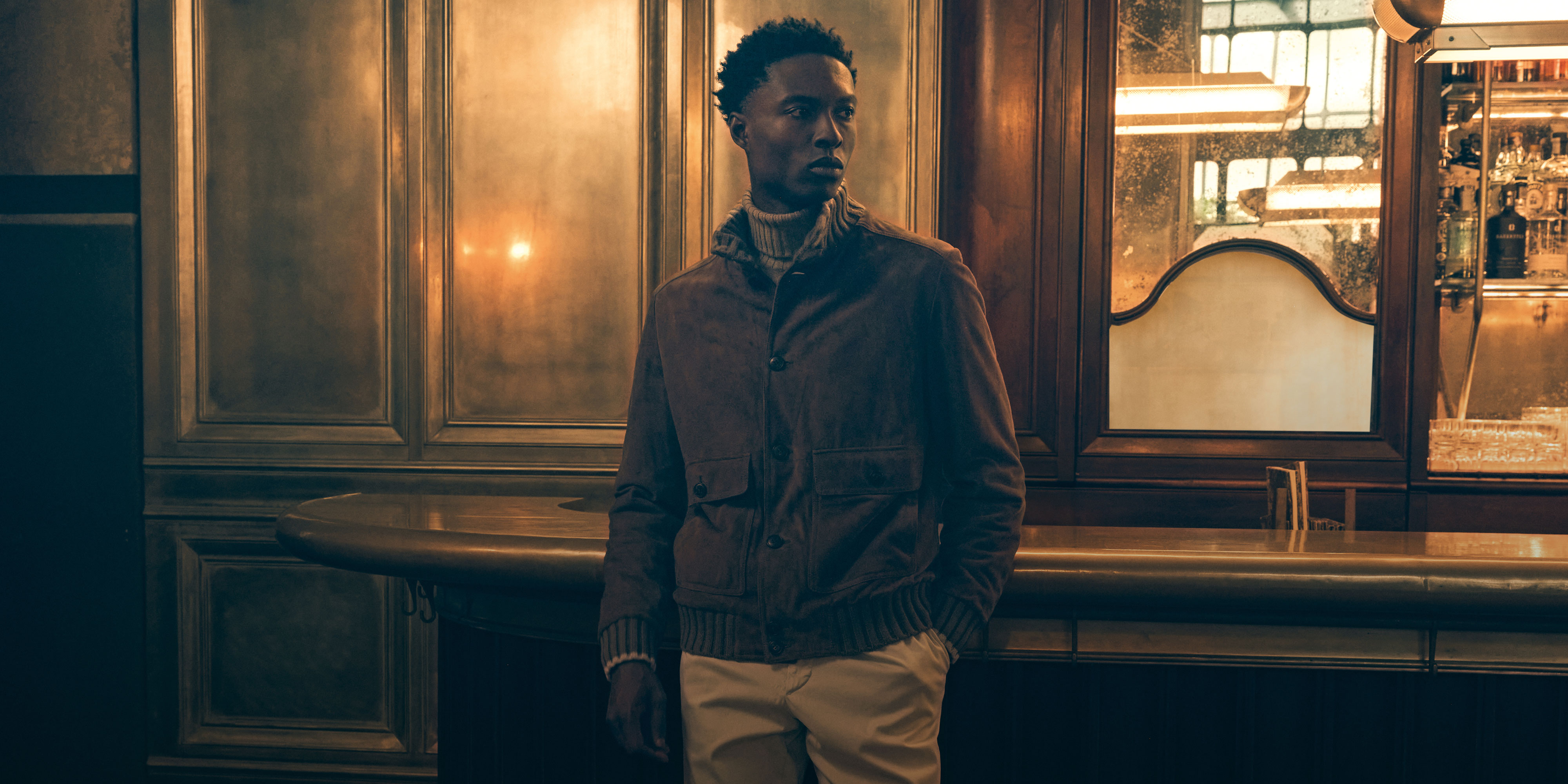 Young man wearing a brown jacket and beige pants standing in a dimly lit vintage bar with wooden panels and a mirror.