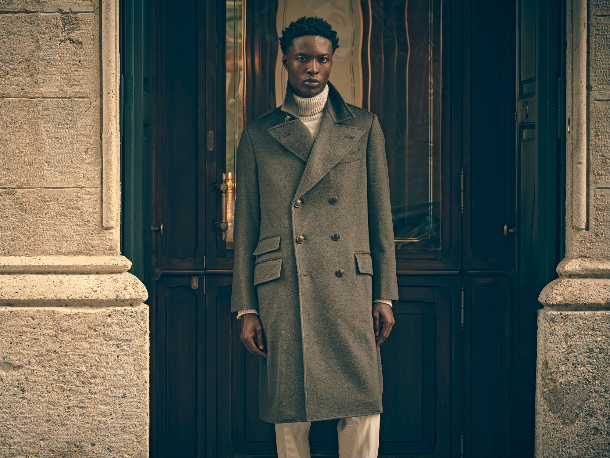 Man wearing a long olive green double-breasted coat and white turtleneck sweater standing in front of a dark wooden door.