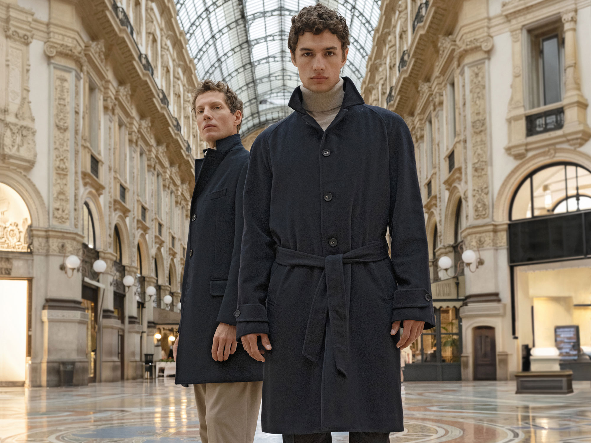 Two men wearing dark navy wool coats standing in an ornate indoor shopping gallery with a glass ceiling.