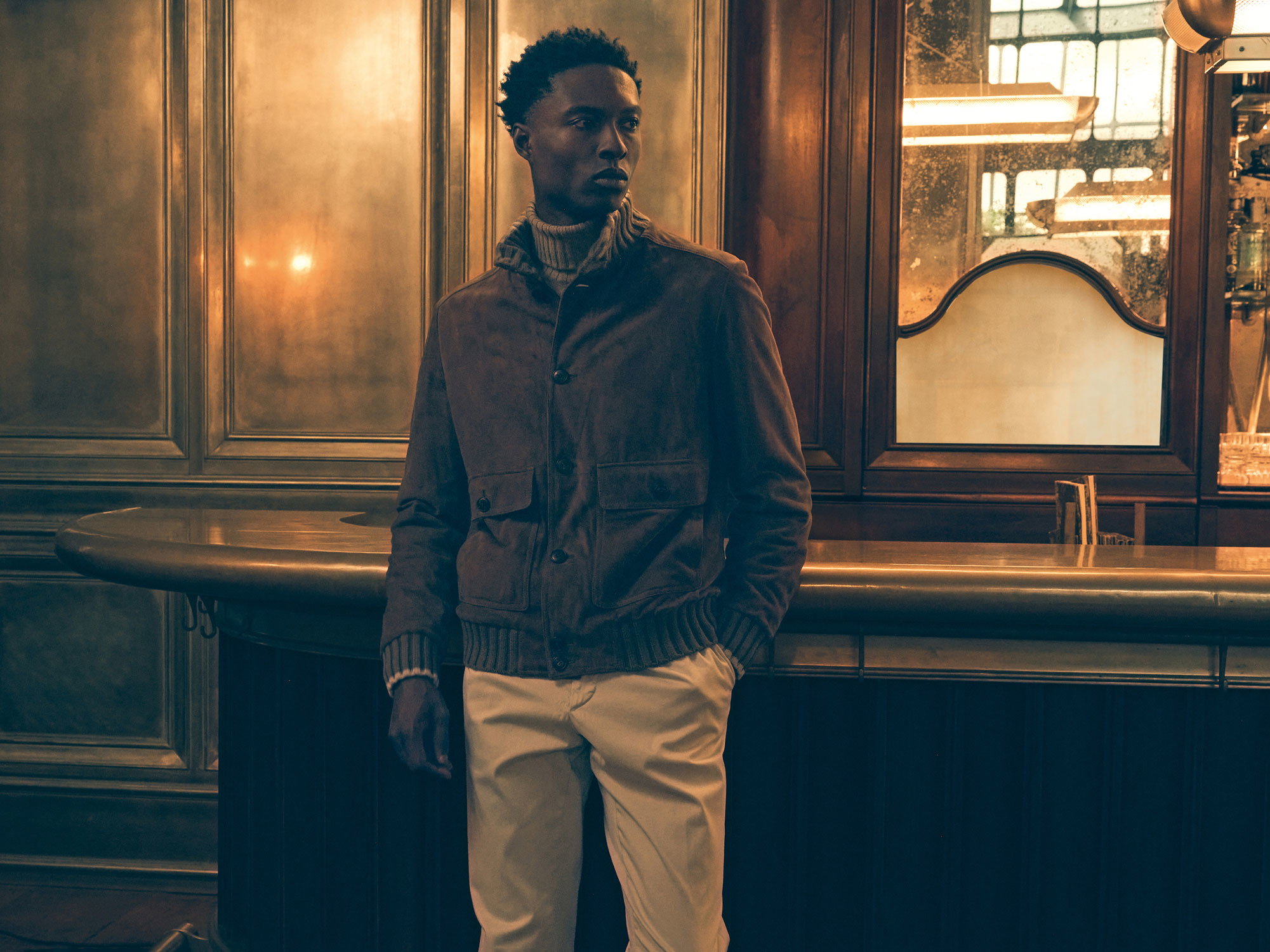 Young man wearing a brown jacket and beige pants leaning against a vintage wooden bar counter in a warmly lit room.