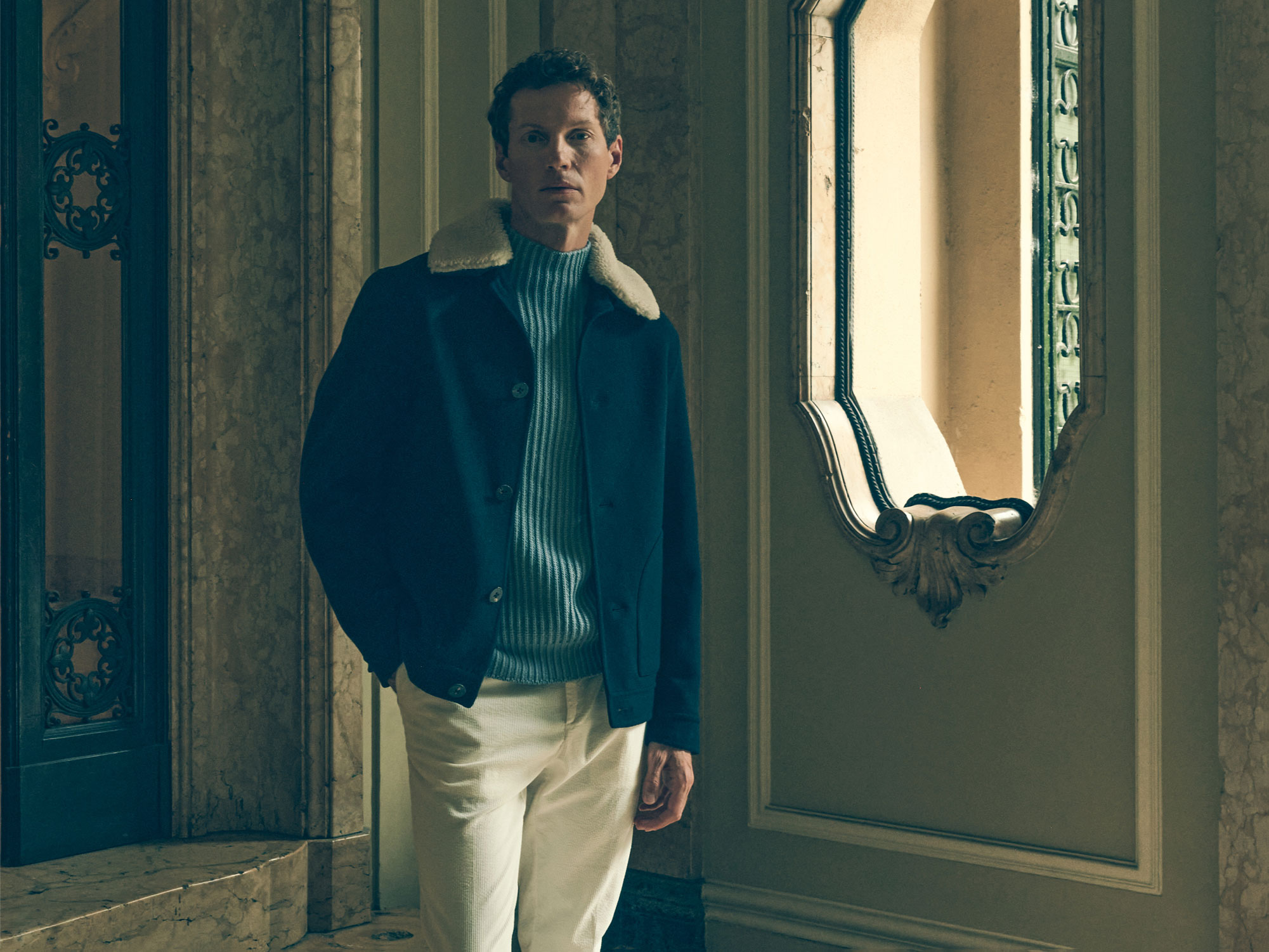 Man wearing a navy blue jacket with white shearling collar and light blue ribbed sweater standing in ornate room near window and mirror.