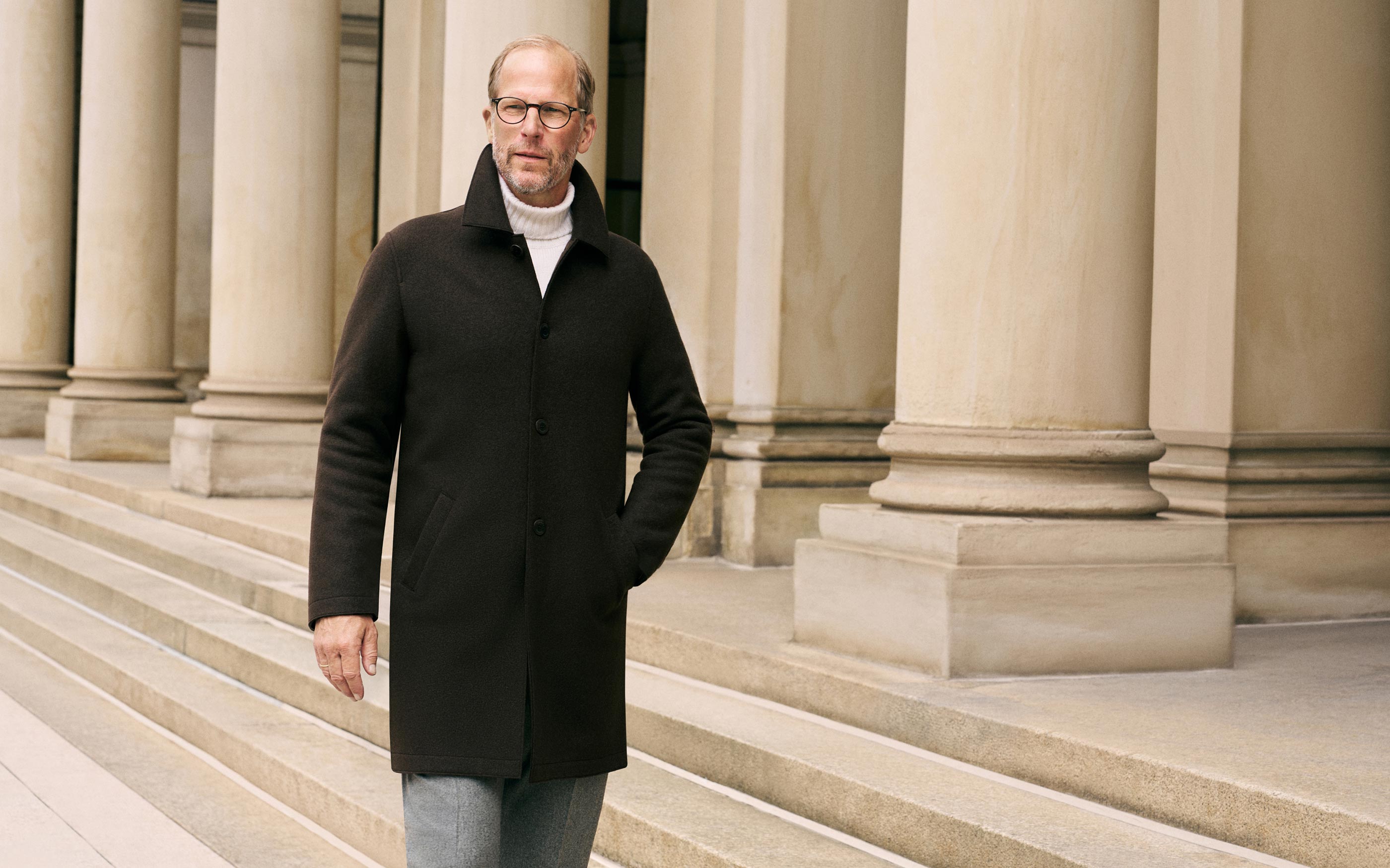 Mann mit Brille trägt braunen Mantel und weißen Rollkragenpullover, geht an großen Sandsteinsäulen und Treppen vorbei.