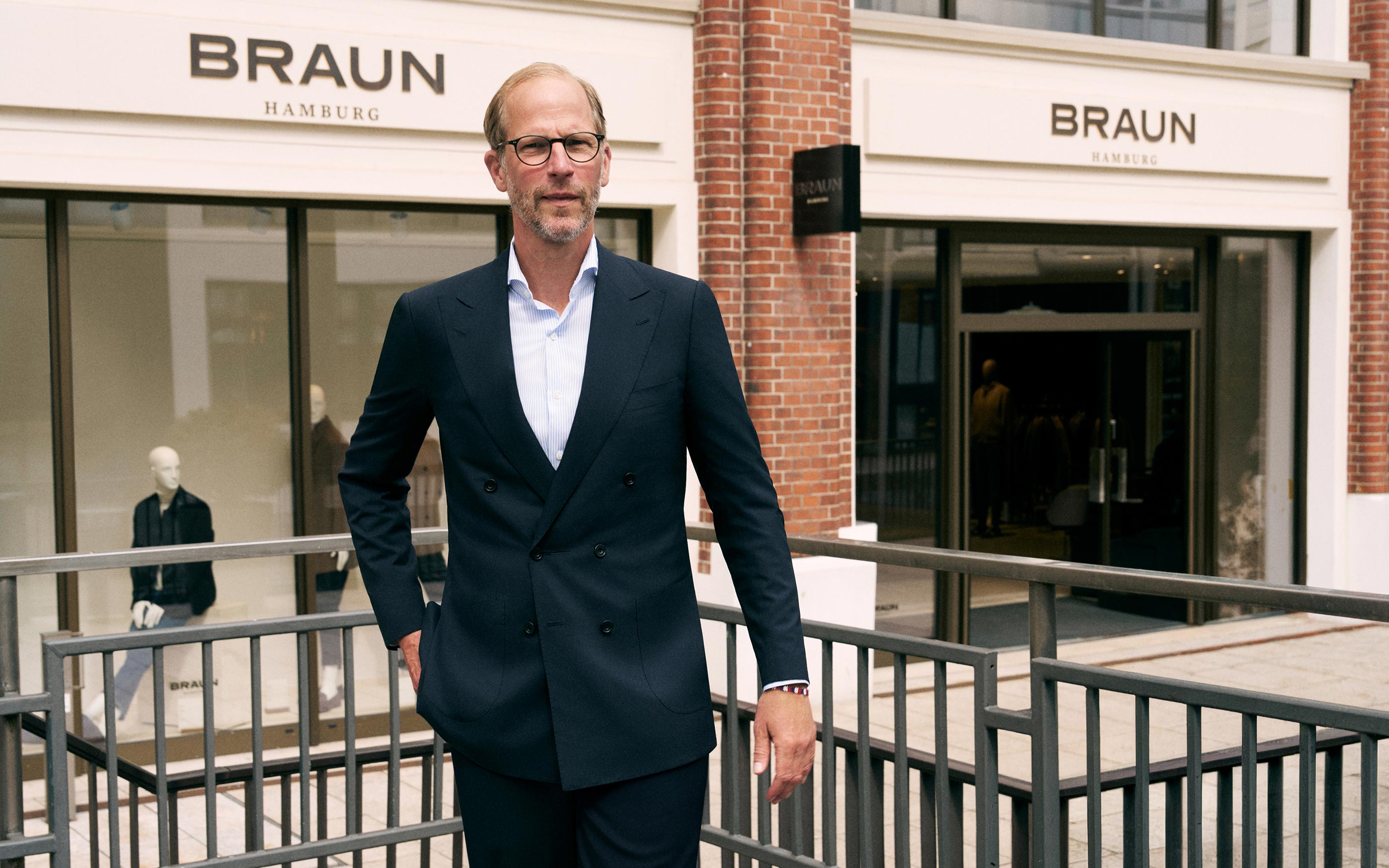 Mann mit Brille und dunkelblauem Anzug steht vor Braun Hamburg Modegeschäft mit Schaufenster und Backsteinfassade.