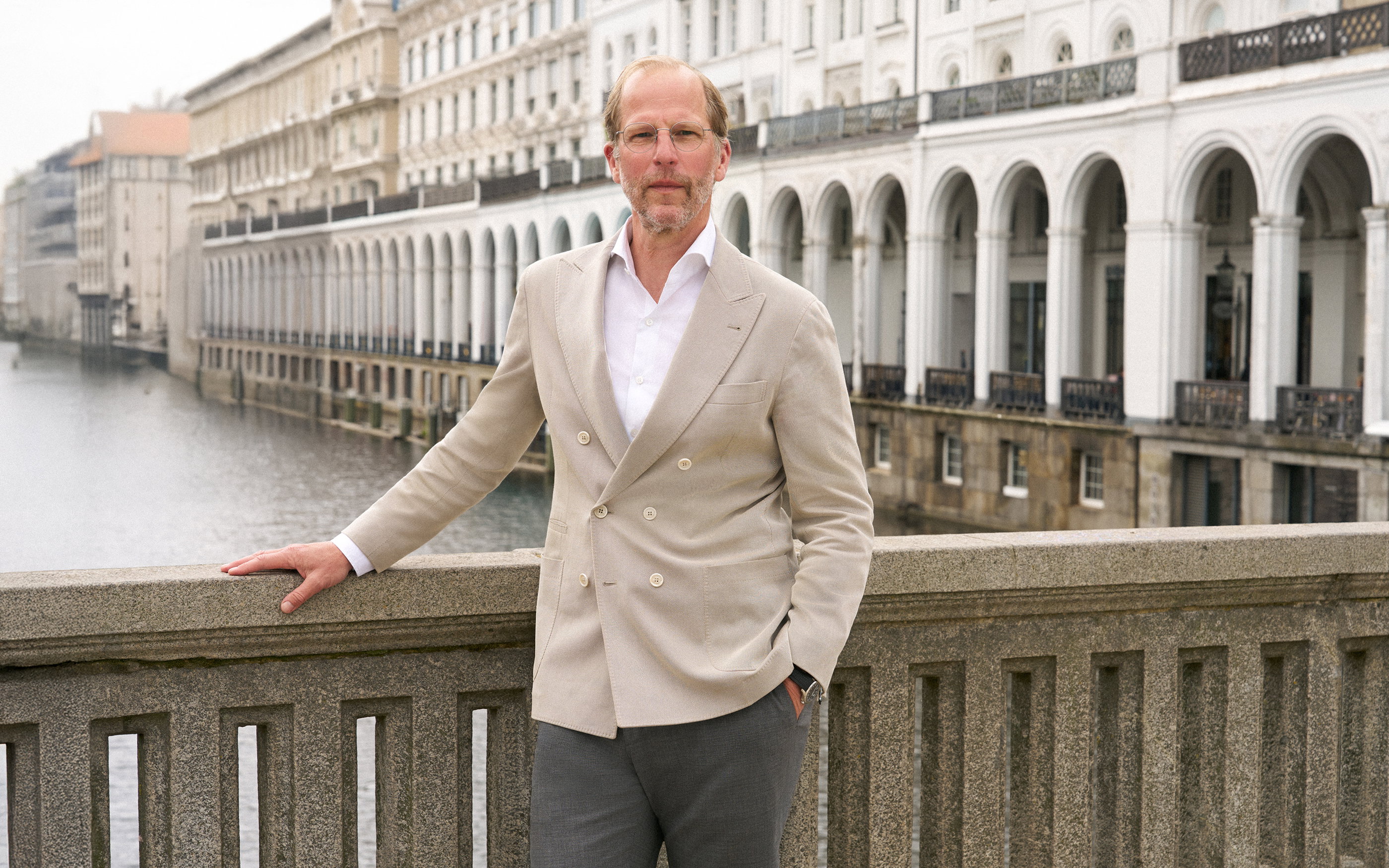 Man in beige double-breasted blazer and glasses standing by stone railing overlooking river and white arched buildings.