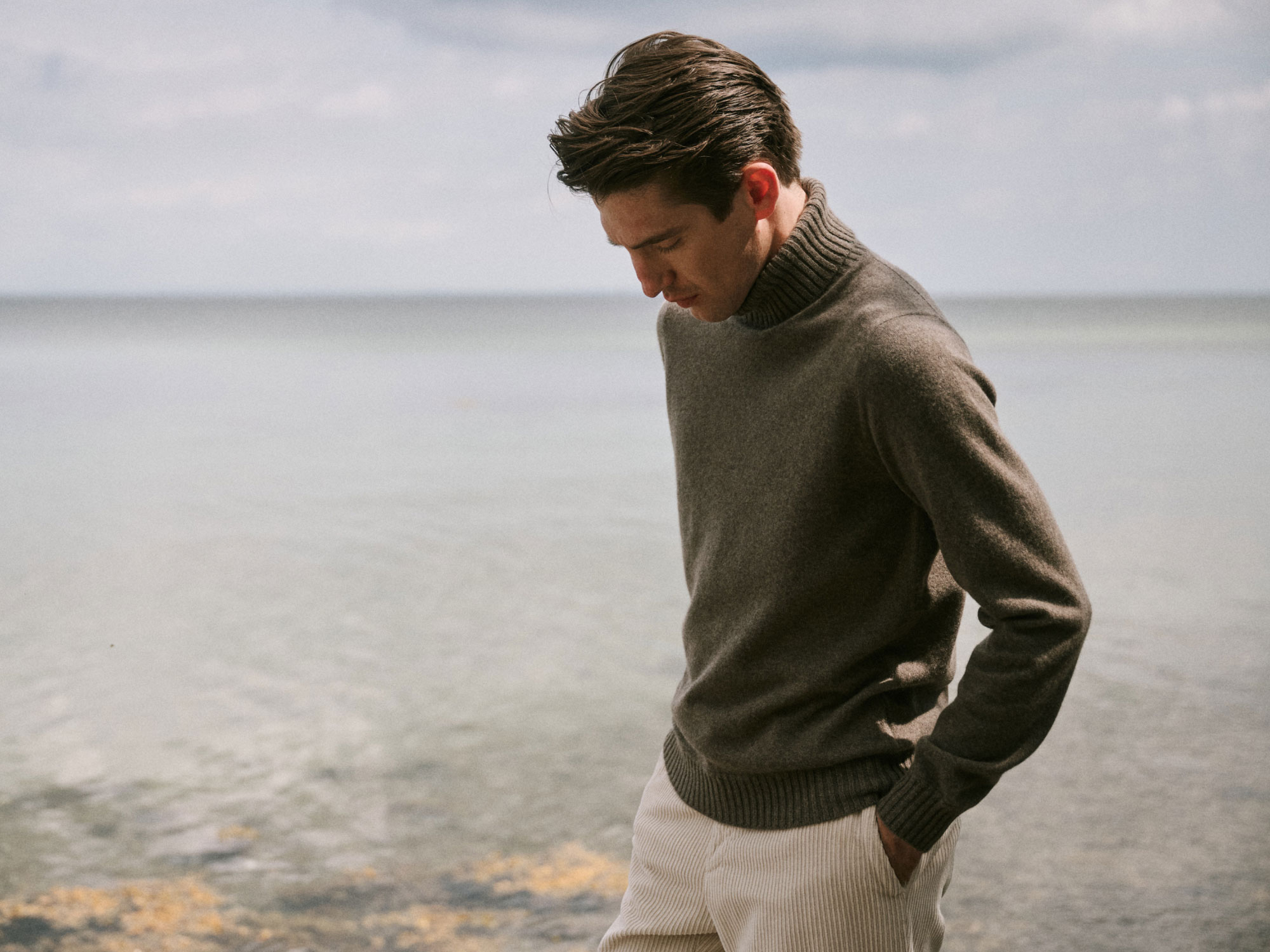 Junger Mann mit braunem Rollkragenpullover und hellem Cordhose steht am Strand und schaut nach unten.