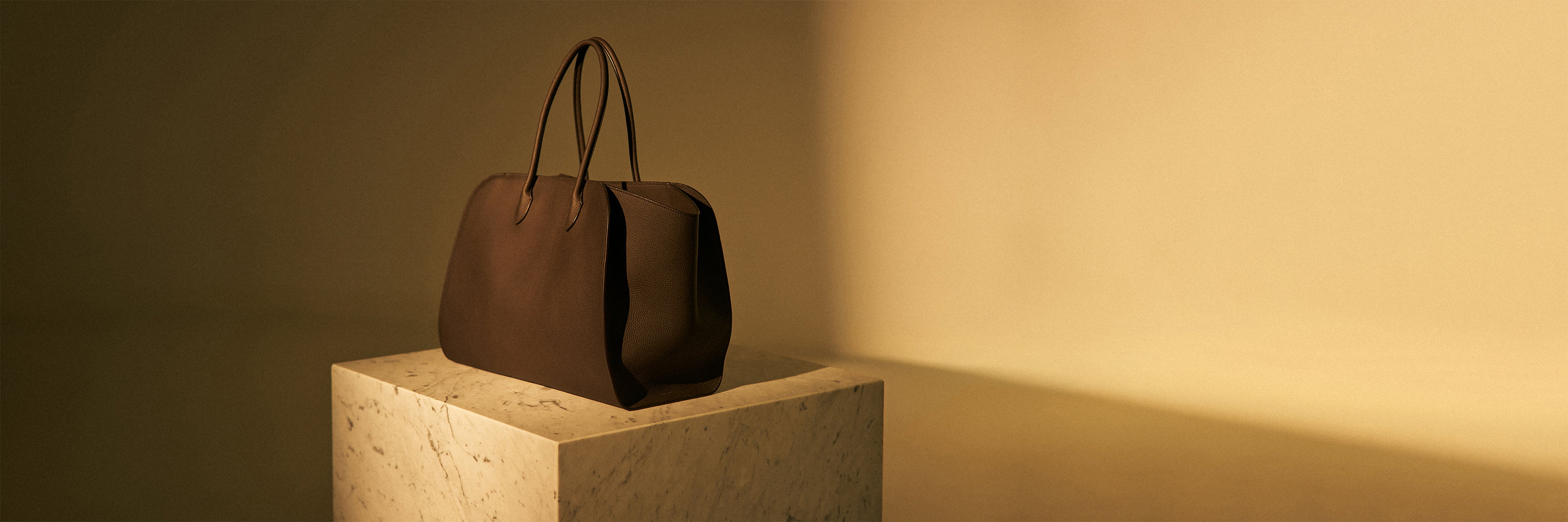 Braune Ledertasche mit langen Henkeln auf einem weißen Marmorsockel vor beige-goldenem Hintergrund.