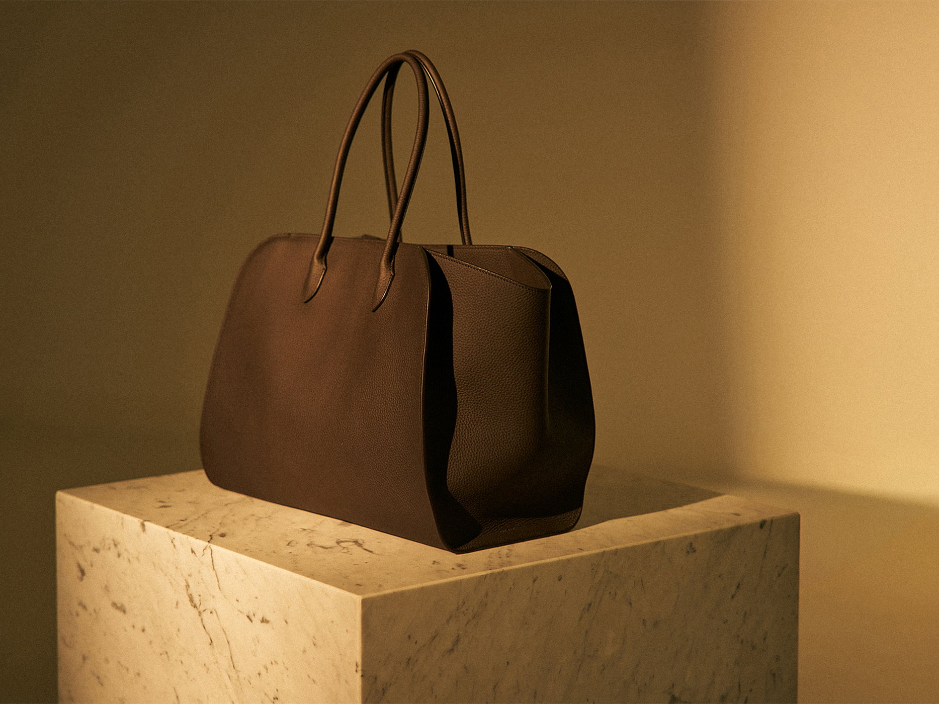 Brown leather handbag with rounded handles placed on a marble pedestal, illuminated by warm, soft light.