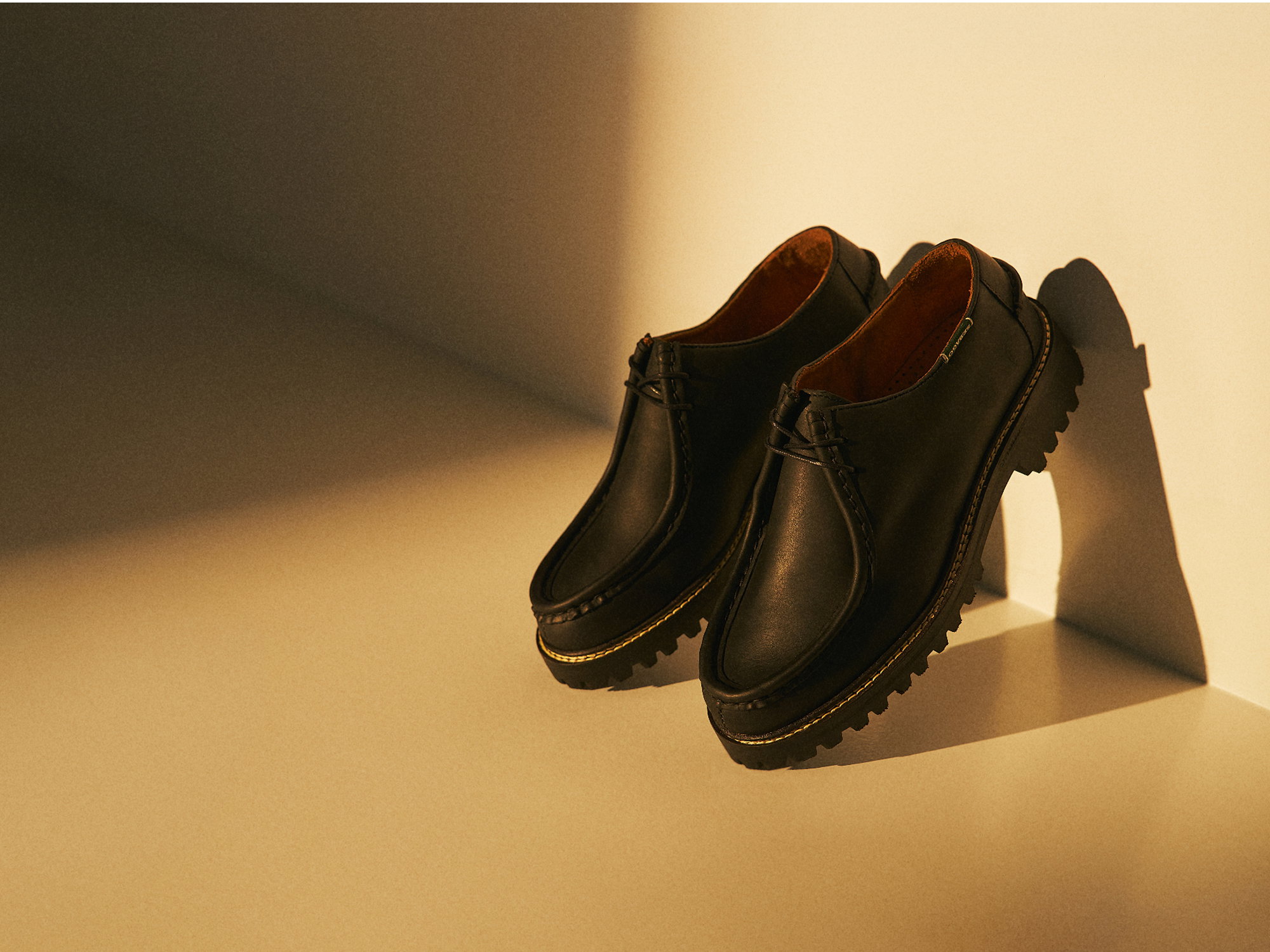 Pair of black leather shoes with rugged soles and yellow stitching, placed on a beige surface with soft shadow.