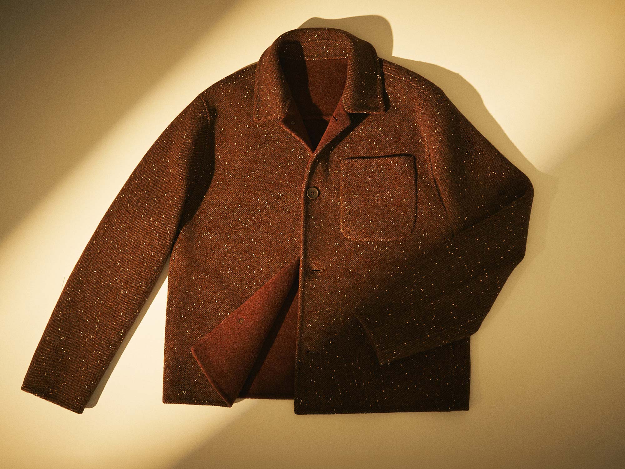 Brown speckled wool jacket with collar and front pocket, laid flat on a beige background with soft lighting and shadow.