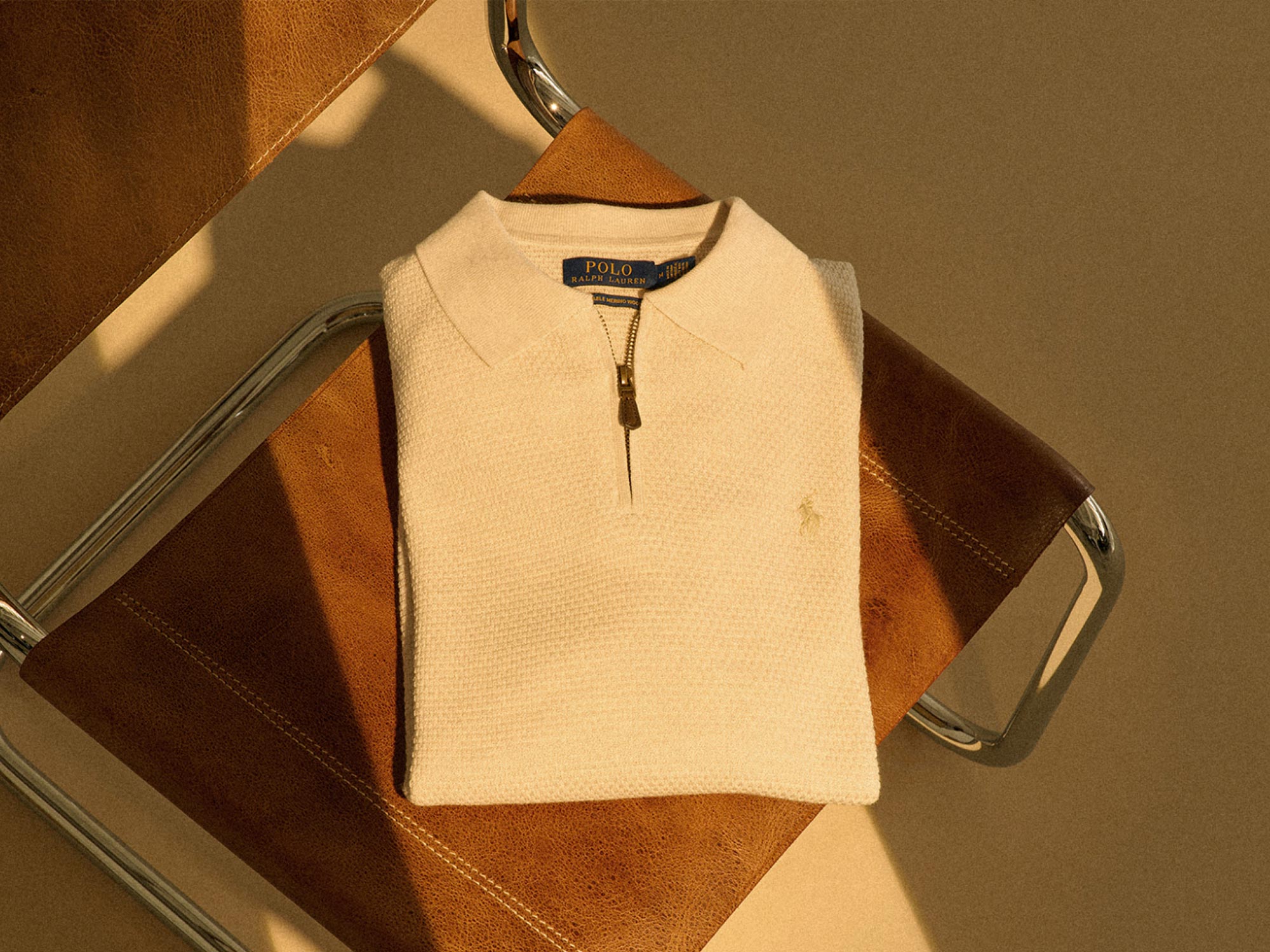 Beige Polo Ralph Lauren Pullover mit Reißverschluss, gefaltet auf braunem Lederstuhl mit Metallrahmen, warme Beleuchtung.