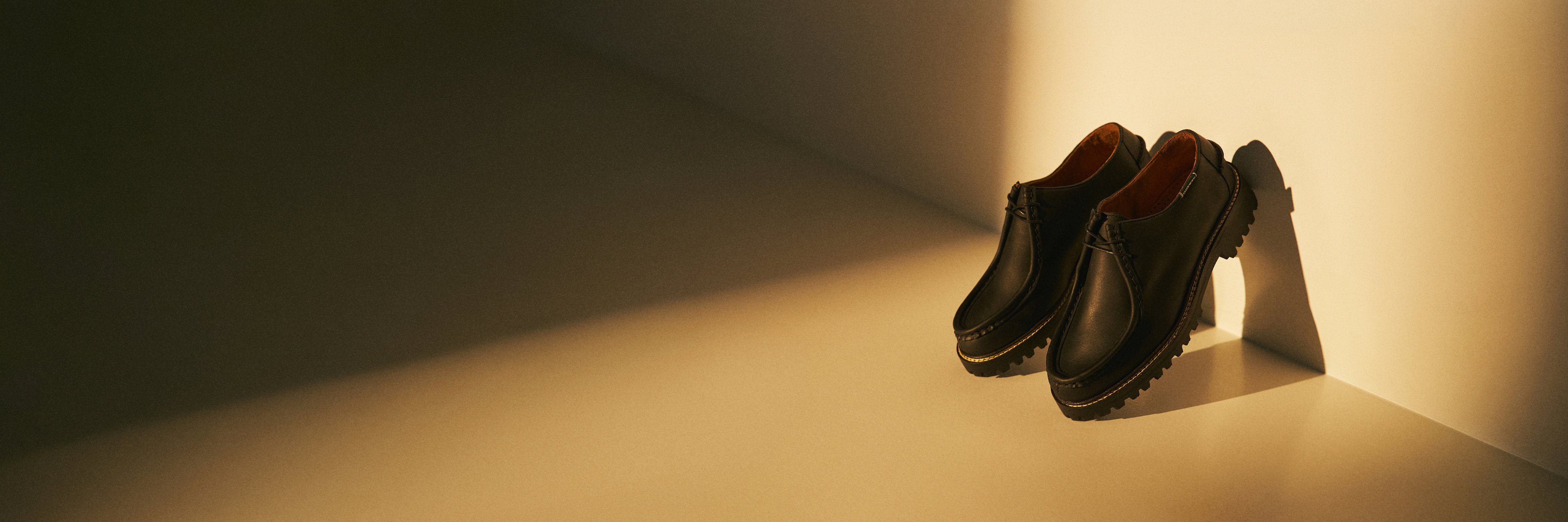 Schwarze Leder-Halbschuhe mit dicker Profilsohle, stehend in einer abgedunkelten Ecke mit warmem Licht und Schatten.