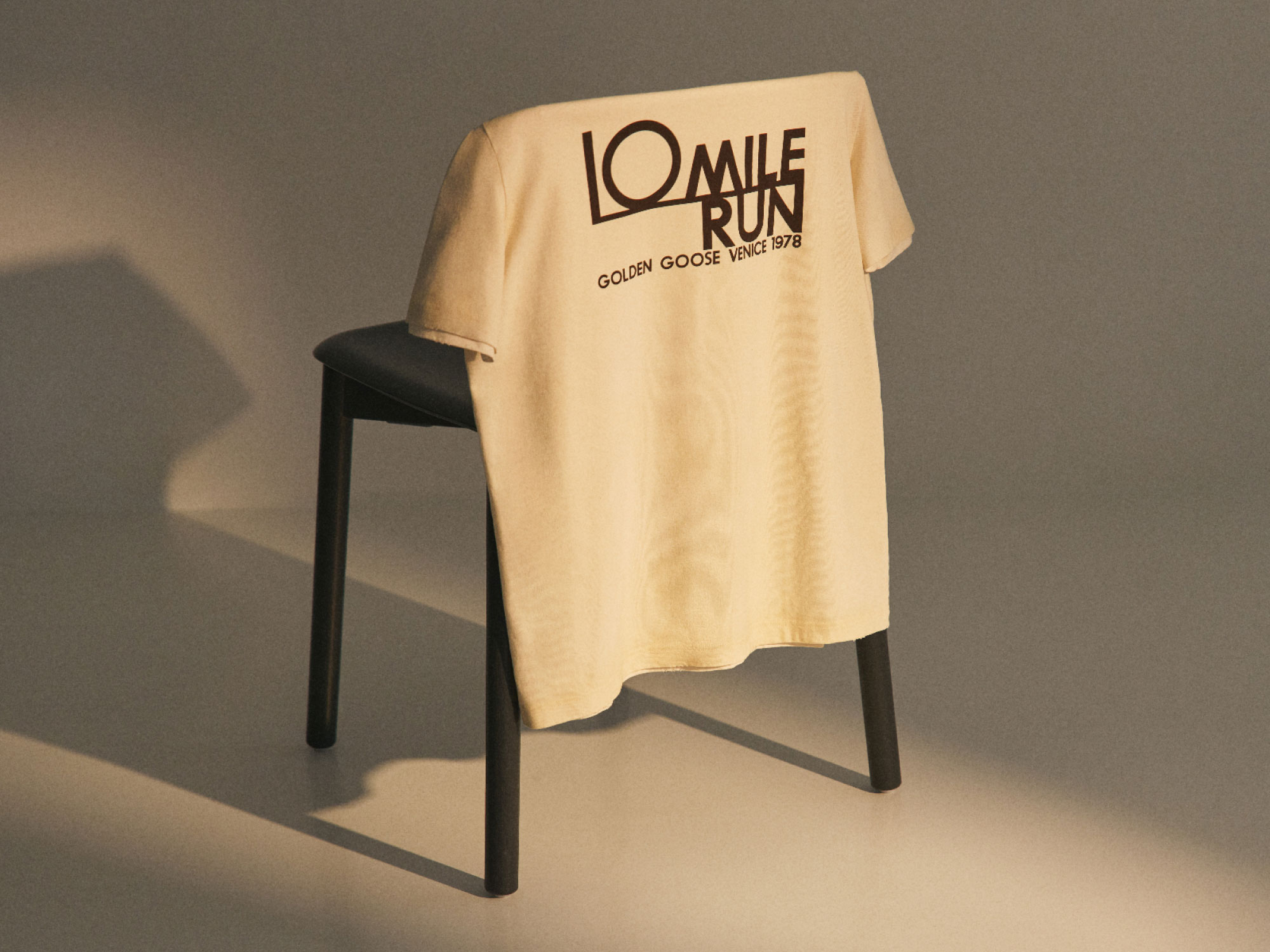 Beige t-shirt with "10 Mile Run Golden Goose Venice 1978" text draped over the back of a black wooden chair casting shadows.