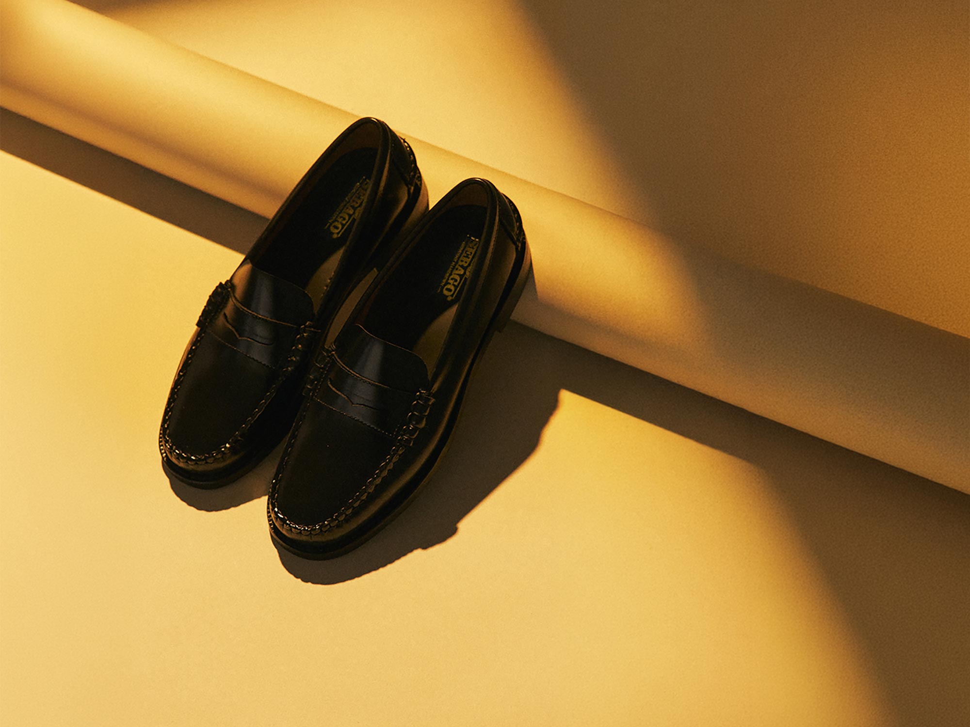 Pair of black leather loafers placed on a yellow surface with soft shadow and warm light highlighting the shoes.