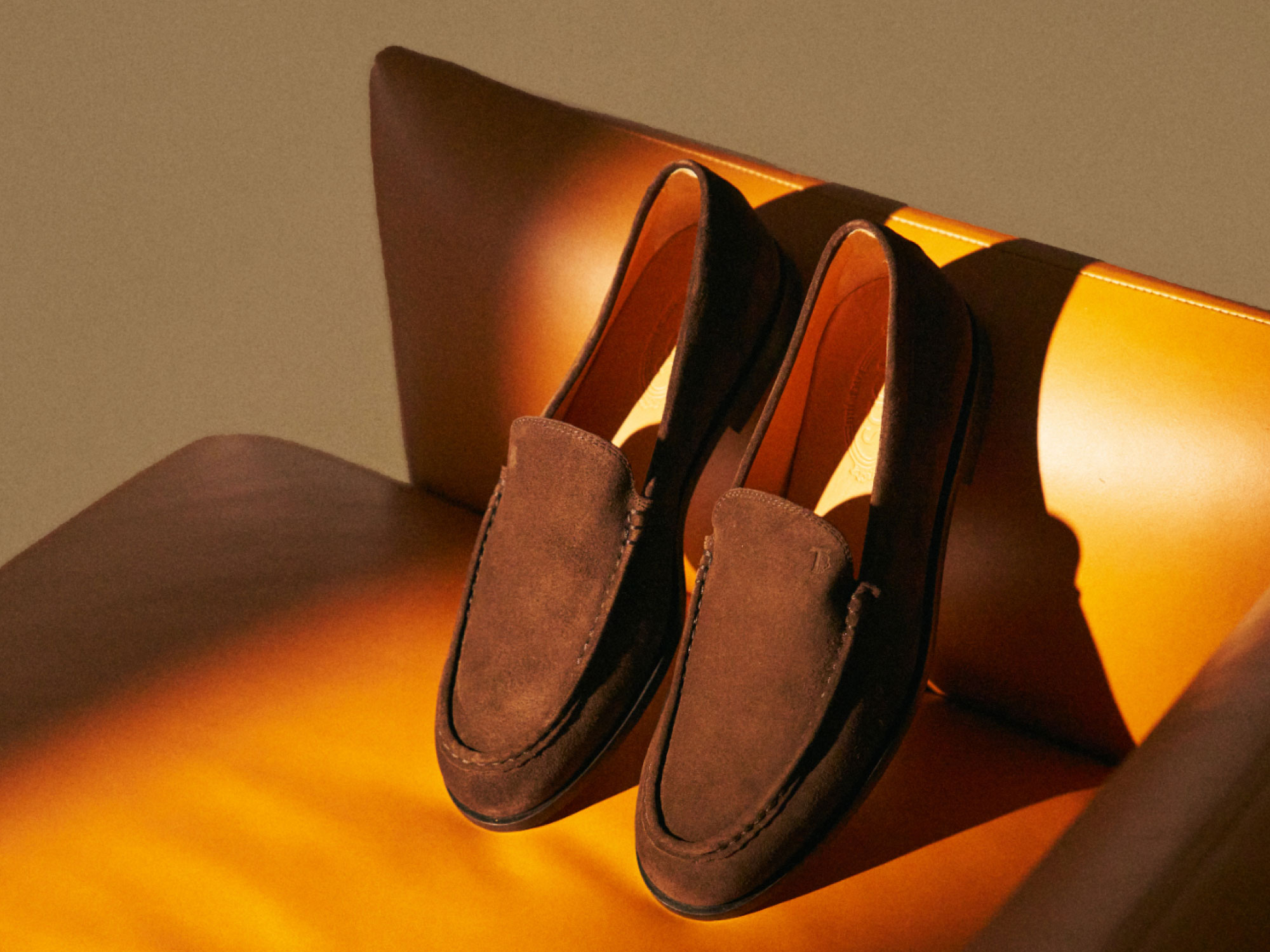 Braune Wildleder-Slipper auf orangefarbenem Ledersessel mit Licht- und Schatteneffekten, Nahaufnahme.