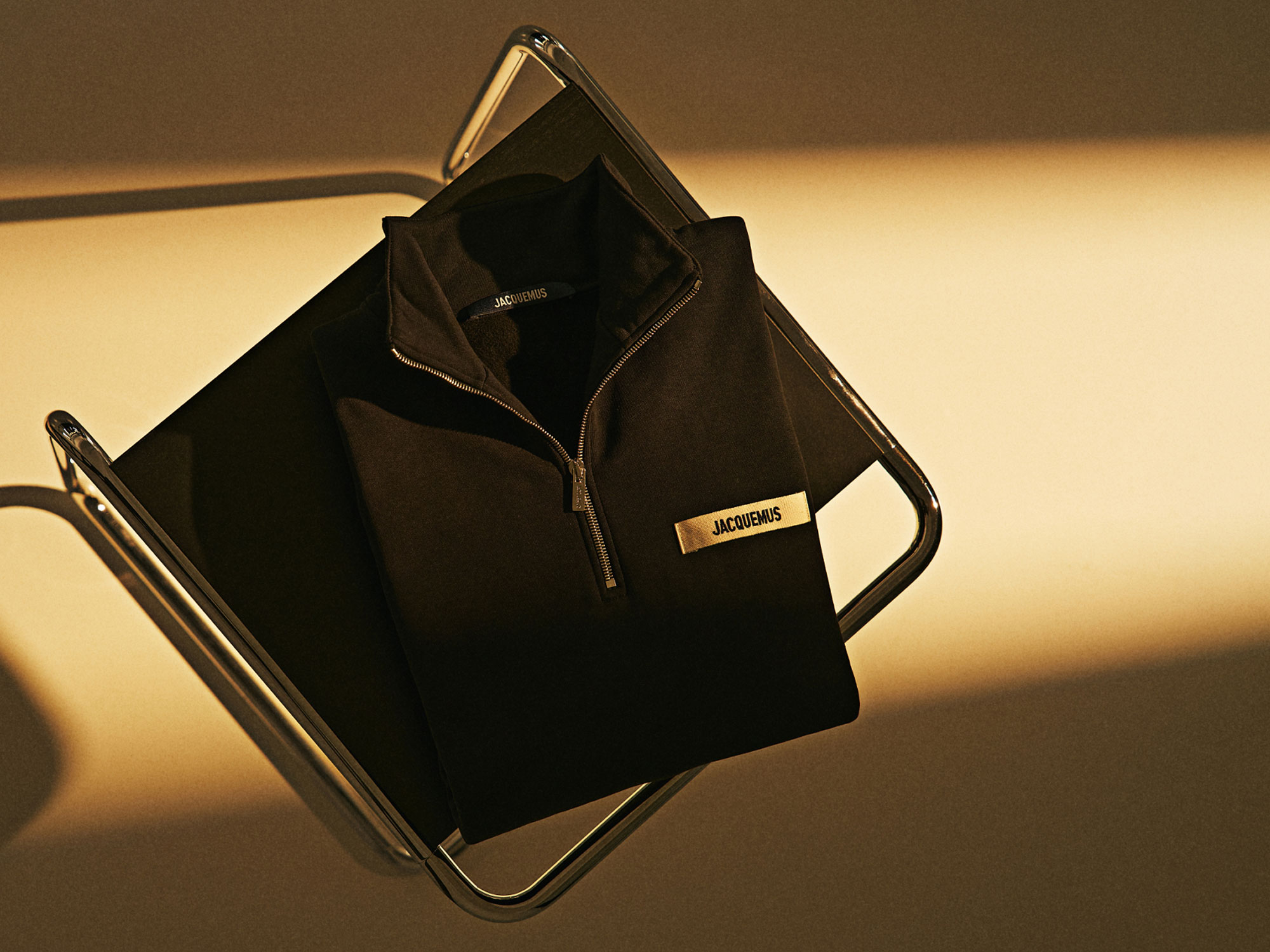 Black Jacquemus zip-up jacket folded on a black chair with chrome frame, warm lighting casting shadows on beige floor.