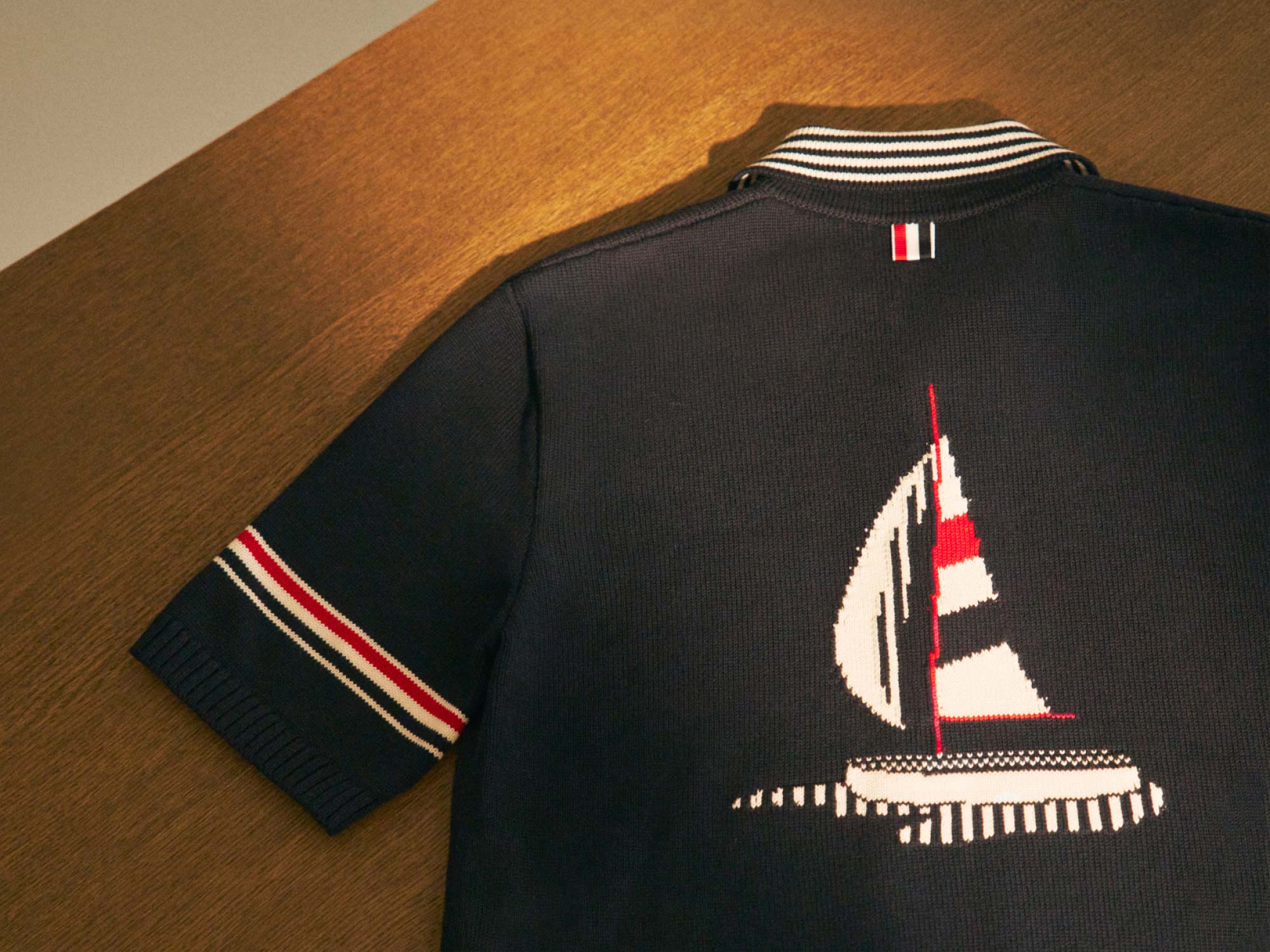 Navy blue knit short-sleeve shirt with striped collar and sleeve cuffs, featuring a red and white sailboat design on wooden surface.