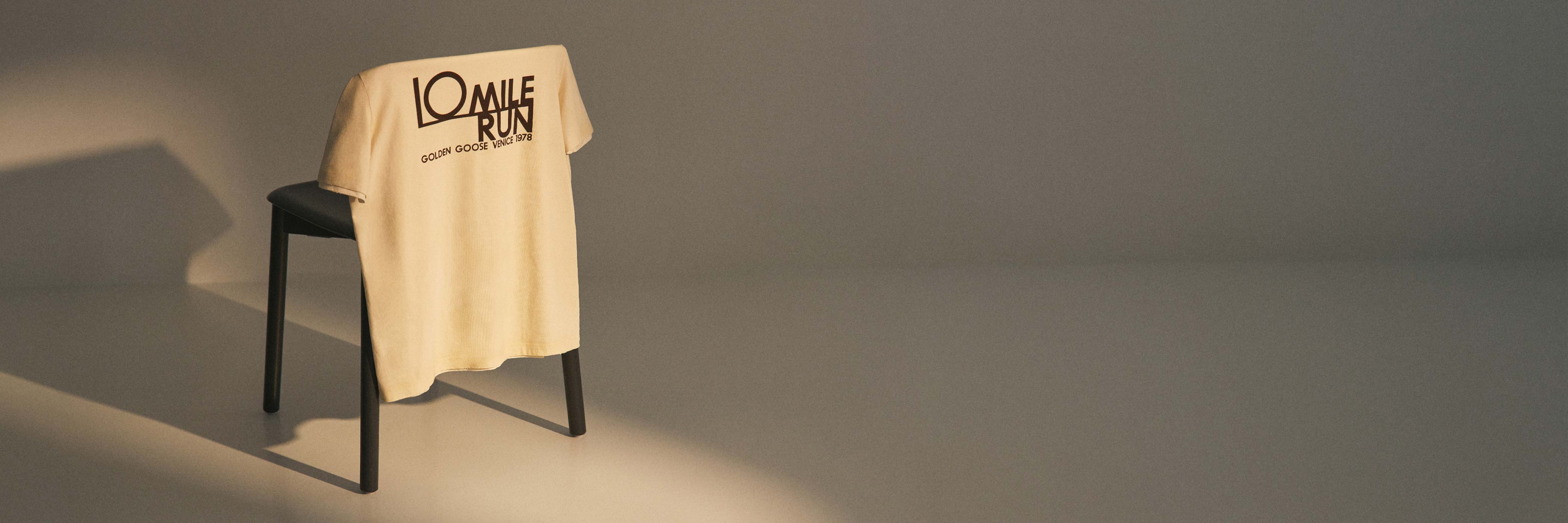 Beiges T-Shirt mit schwarzem Aufdruck "10 Mile Run Golden Goose Venice 1978" hängt über eine schwarze Stuhllehne in minimalistischem Raum.