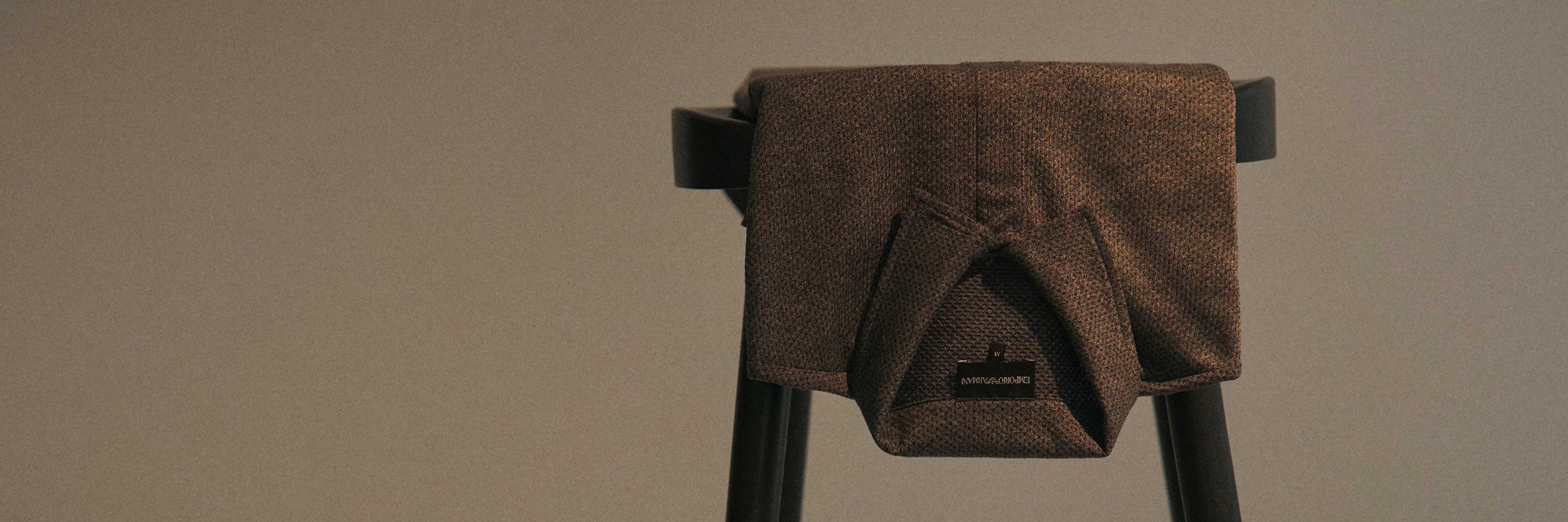 Brauner, fein strukturierter Pullover mit Armani-Label, lässig über die Lehne eines schwarzen Stuhls gelegt.