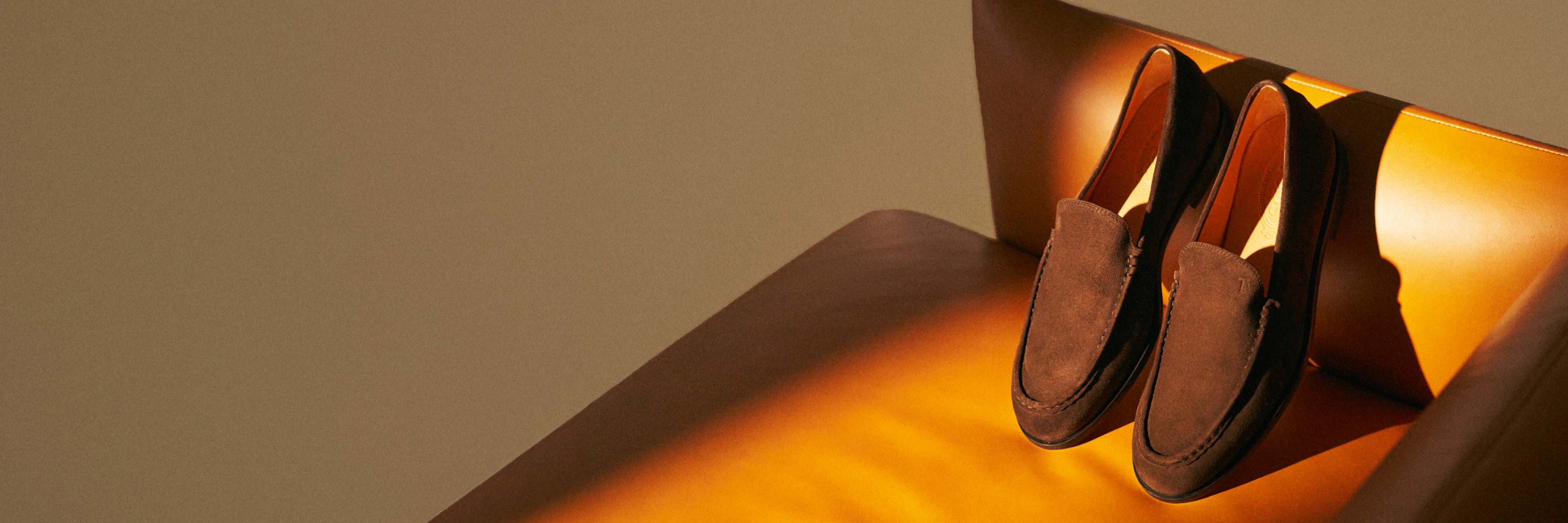 Braune Wildleder-Herrenschuhe liegen nebeneinander auf einem orangefarbenen Ledersessel mit warmem Licht.