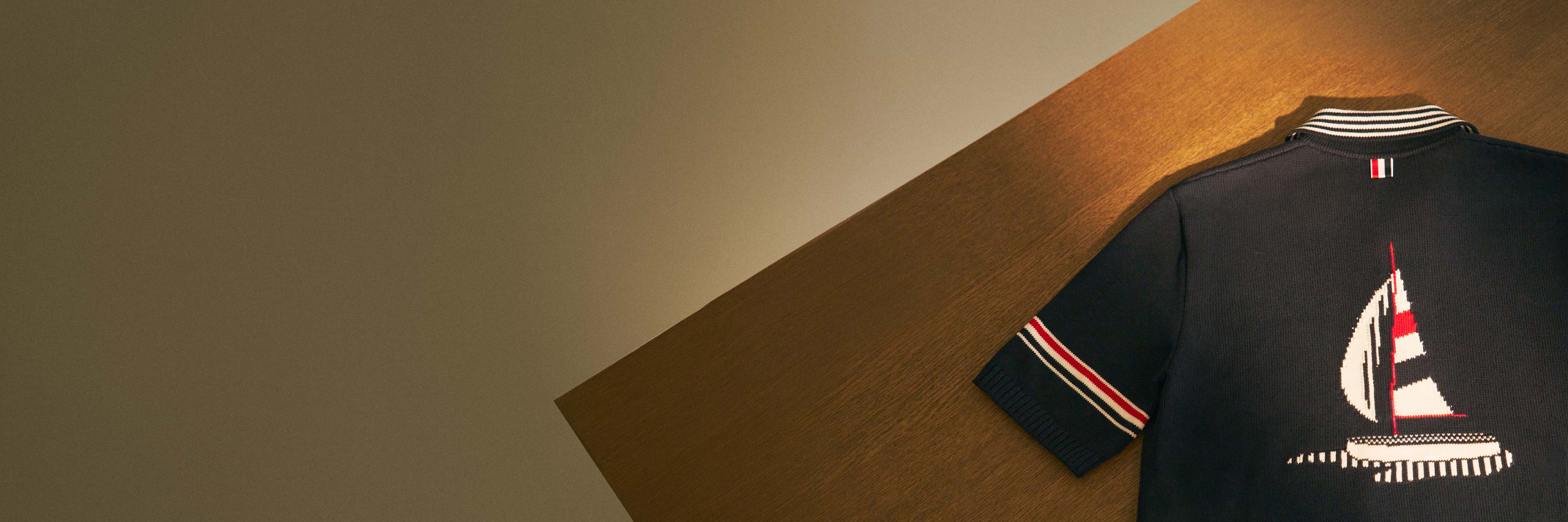 Dunkelblaues Poloshirt mit weiß-rotem Segelboot-Motiv, gestreiftem Kragen und Ärmeln, auf Holztisch liegend.