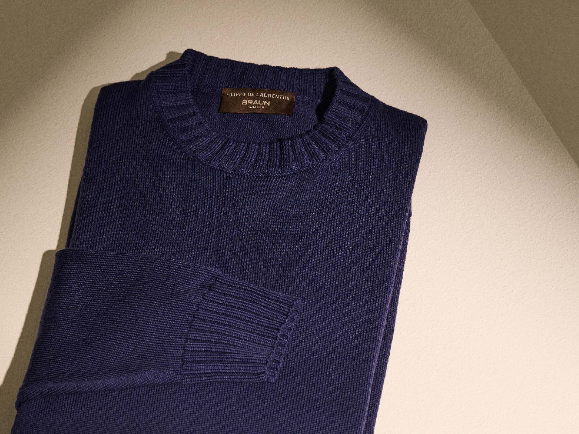 Folded navy blue ribbed knit sweater with a round neck and a visible brown label reading "Filippo De Laurentiis Braun Hamburg" on beige background.