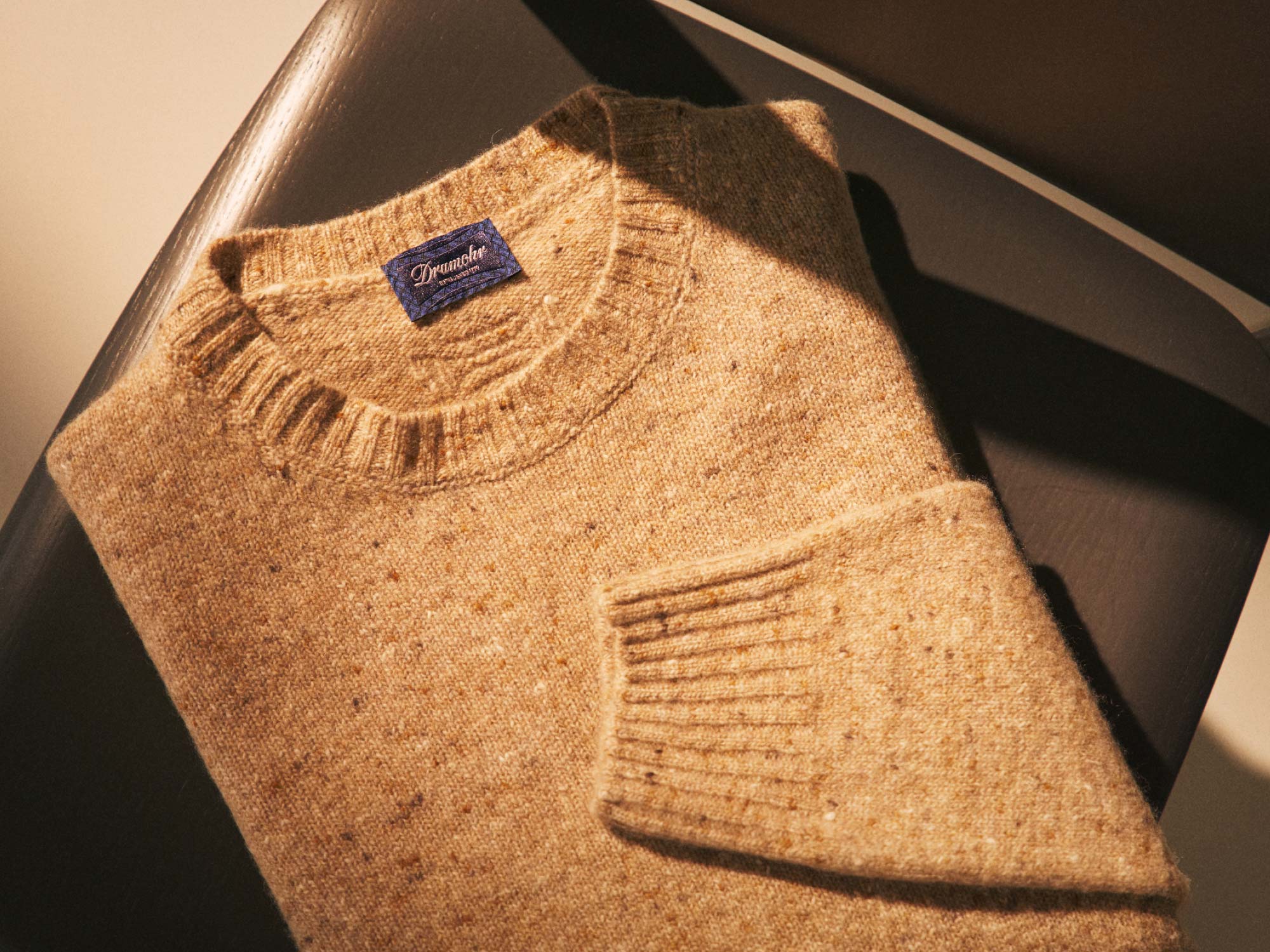 Folded beige knit sweater with ribbed collar and cuffs, featuring a dark blue "Drumohr" label on a dark brown surface.