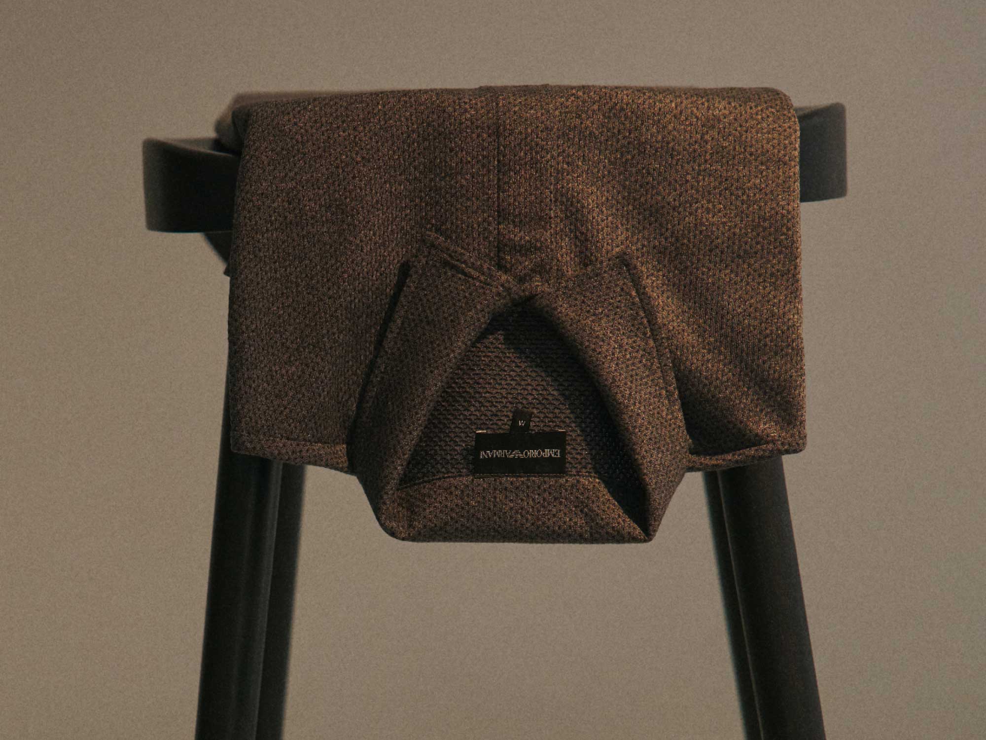 Brown textured knit sweater draped over the backrest of a black chair against a plain beige wall.