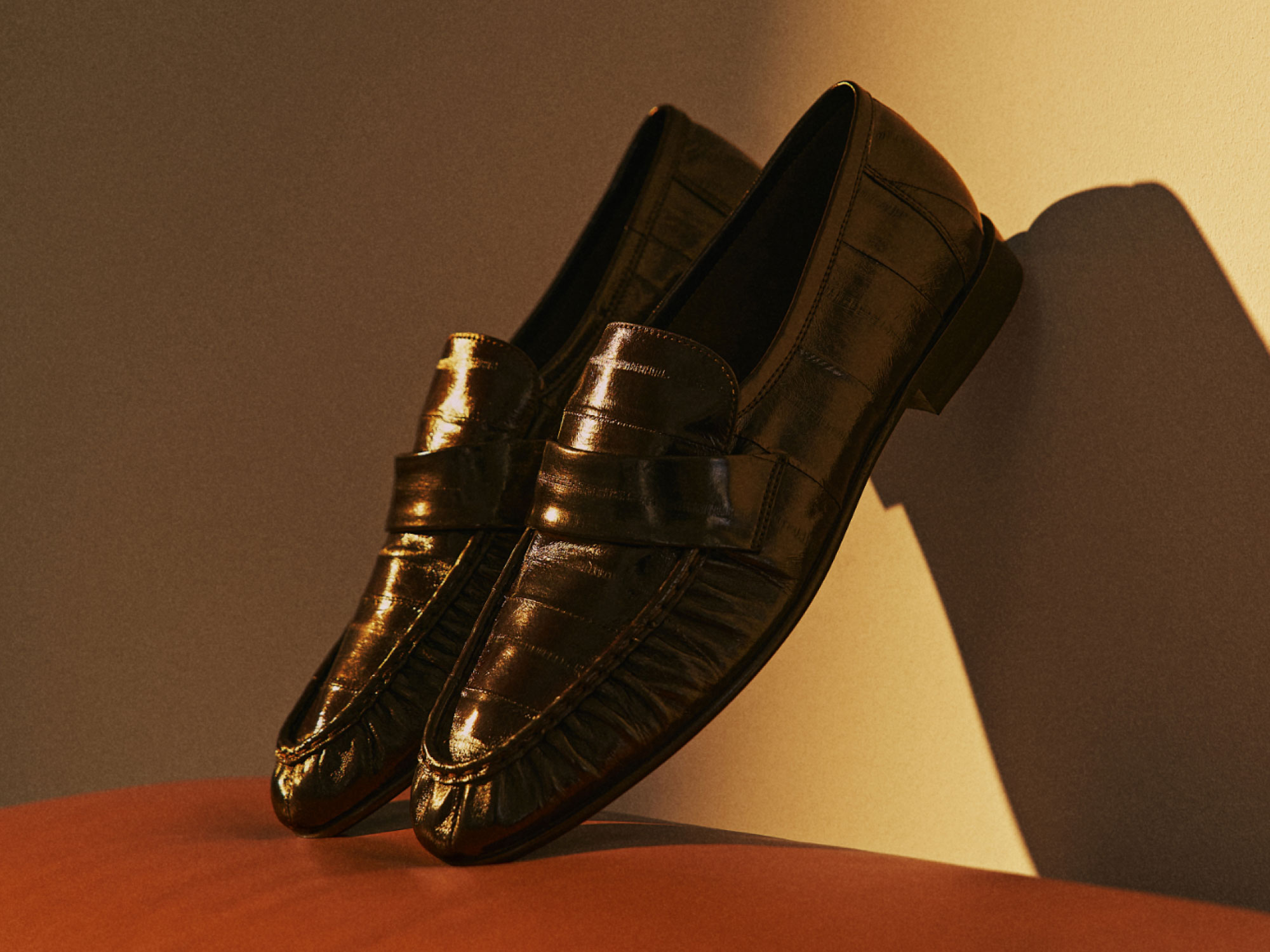 Braune Leder-Loafer mit Kroko-Prägung, auf orangefarbenem Sitzmöbel vor beiger Wand stehend mit Schattenwurf.