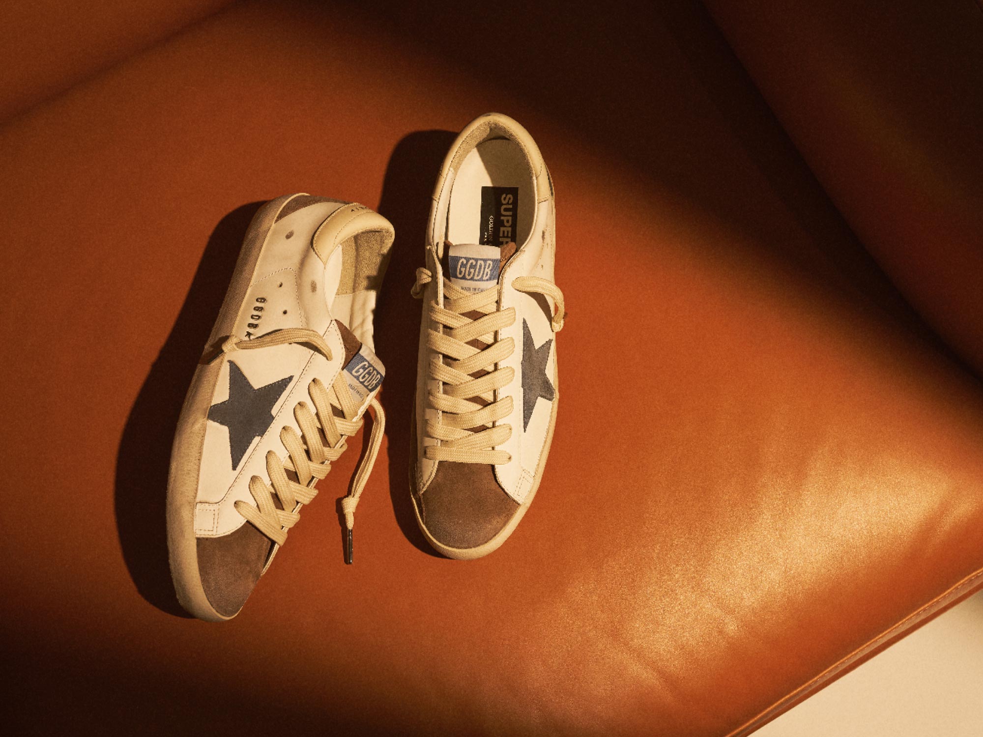 Weiße Sneaker mit dunkelblauem Stern, braunen Akzenten und beigen Schnürsenkeln liegen auf braunem Ledersessel.