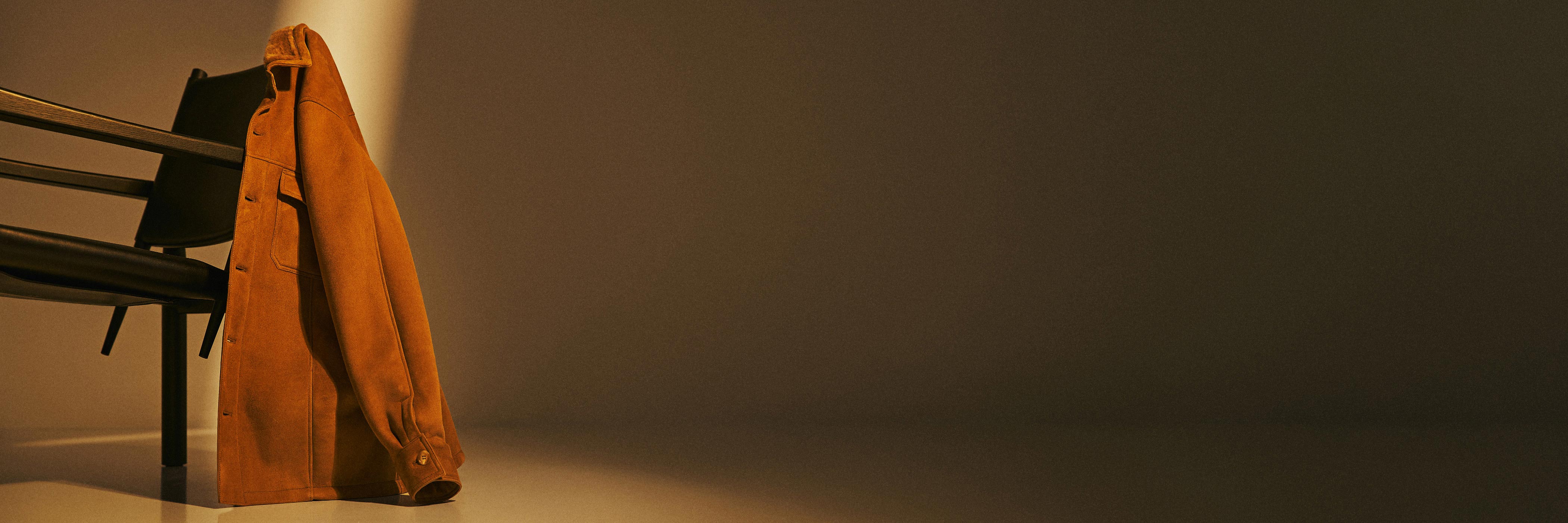 Braune Wildlederjacke hängt auf der Rückenlehne eines schwarzen Stuhls, warme Lichtstimmung, minimalistische Szene.