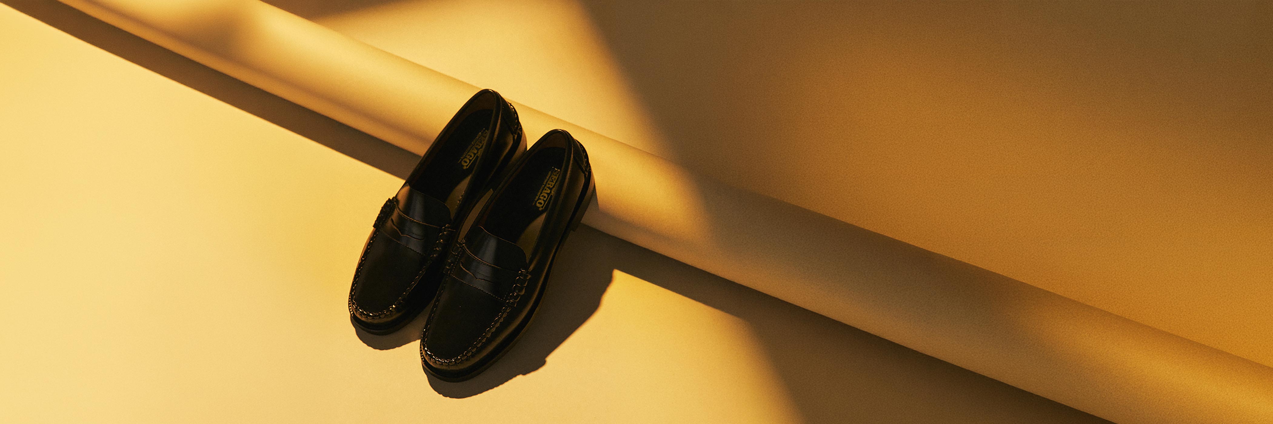 Schwarze Lackleder-Loafers auf gelbem Untergrund mit Schatten und gerolltem Papier im Hintergrund.