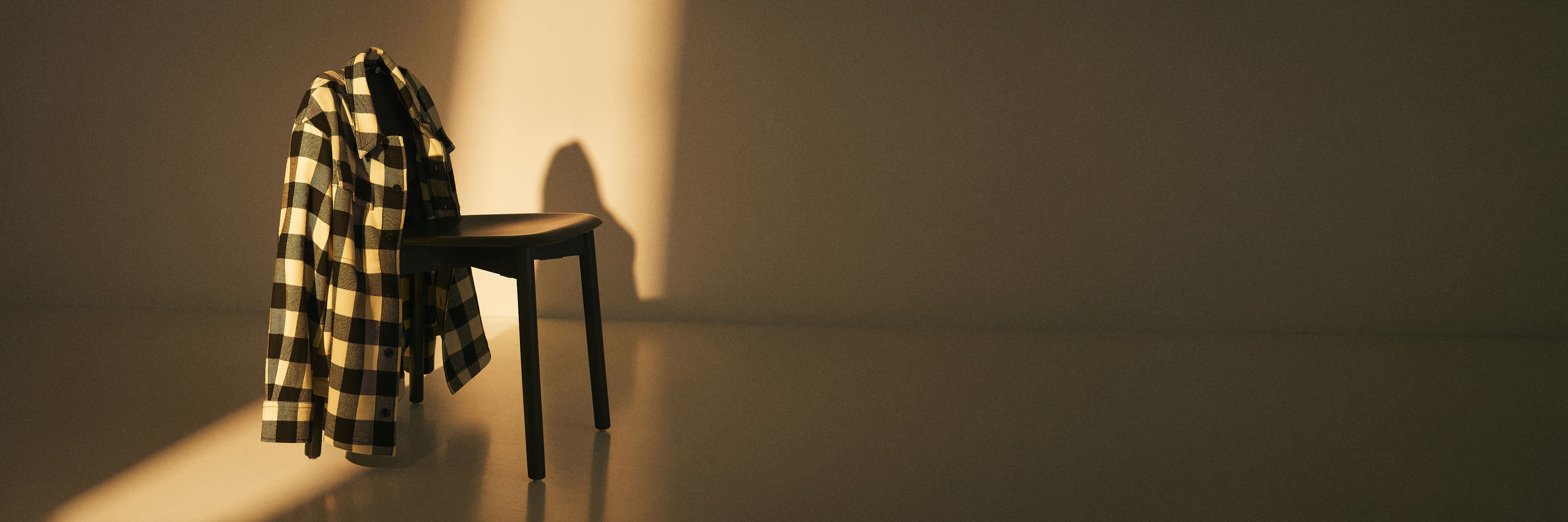 Schwarzer Stuhl mit schwarz-weiß kariertem Hemd, warmes Licht fällt schräg auf Stuhl und Wand im minimalistischen Raum.